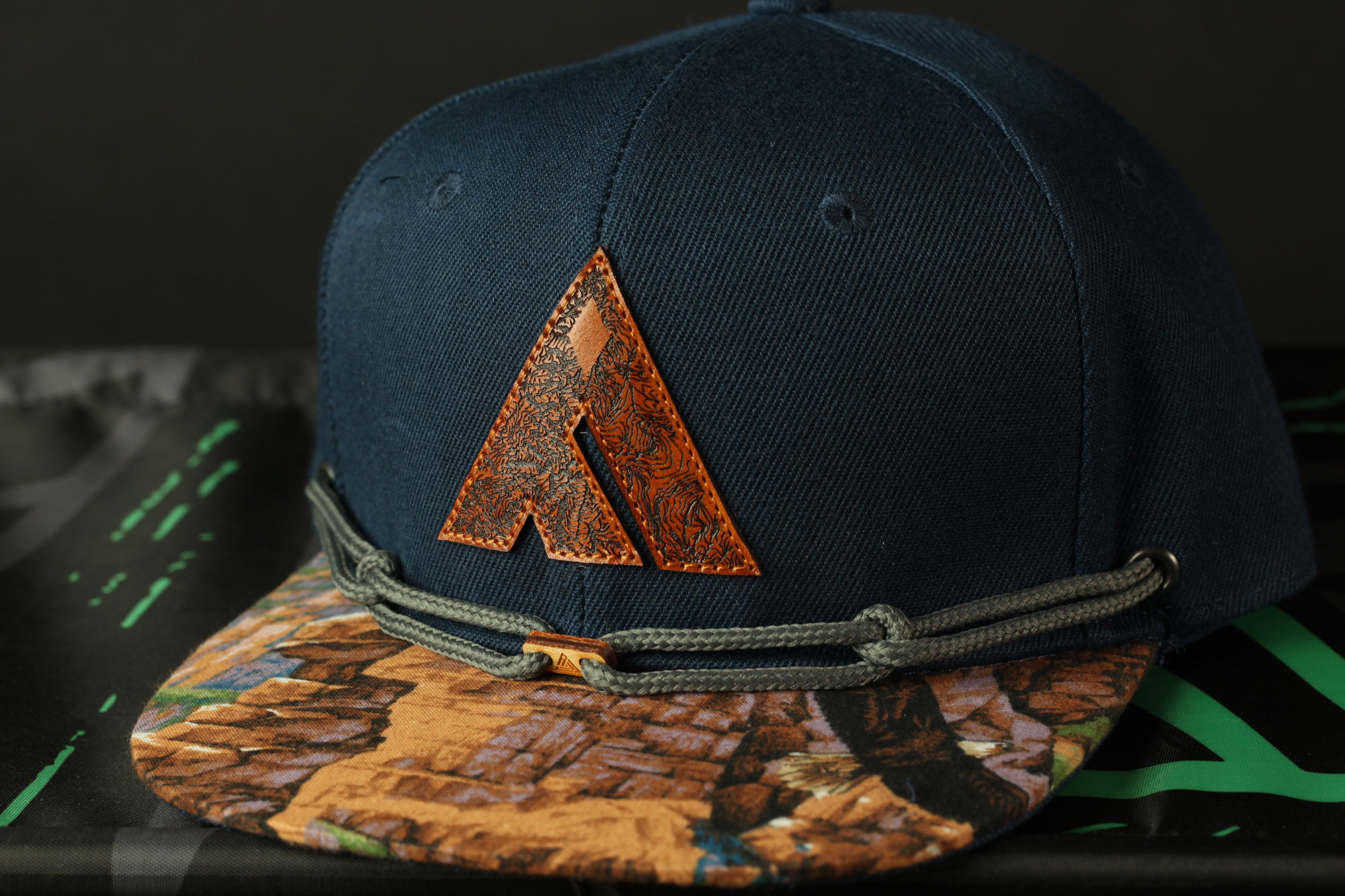A navy blue baseball cap with a brown leather logo on the front, resting on a black surface with a green and black patterned background.