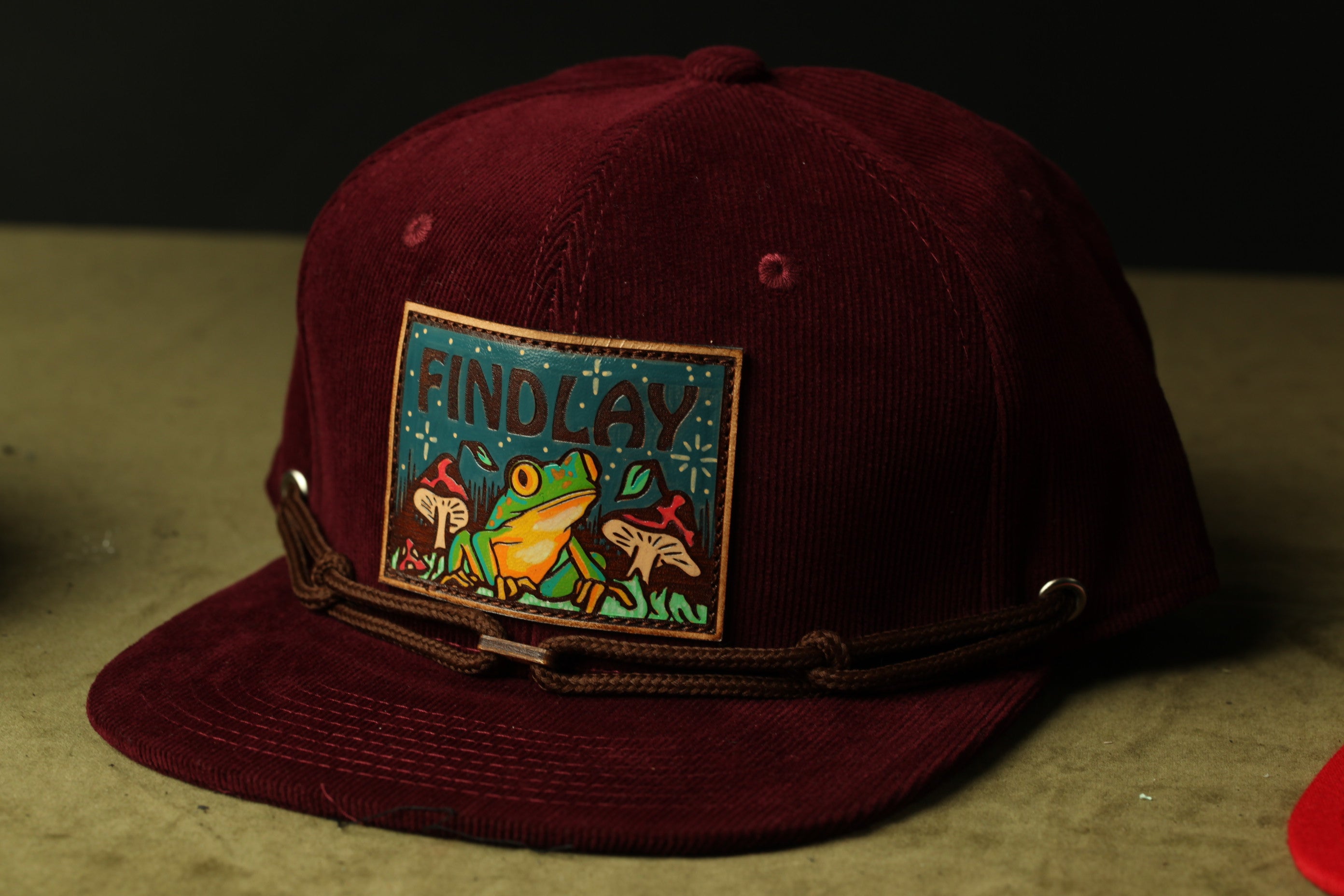 A burgundy baseball cap with a patch featuring a frog and mushrooms.