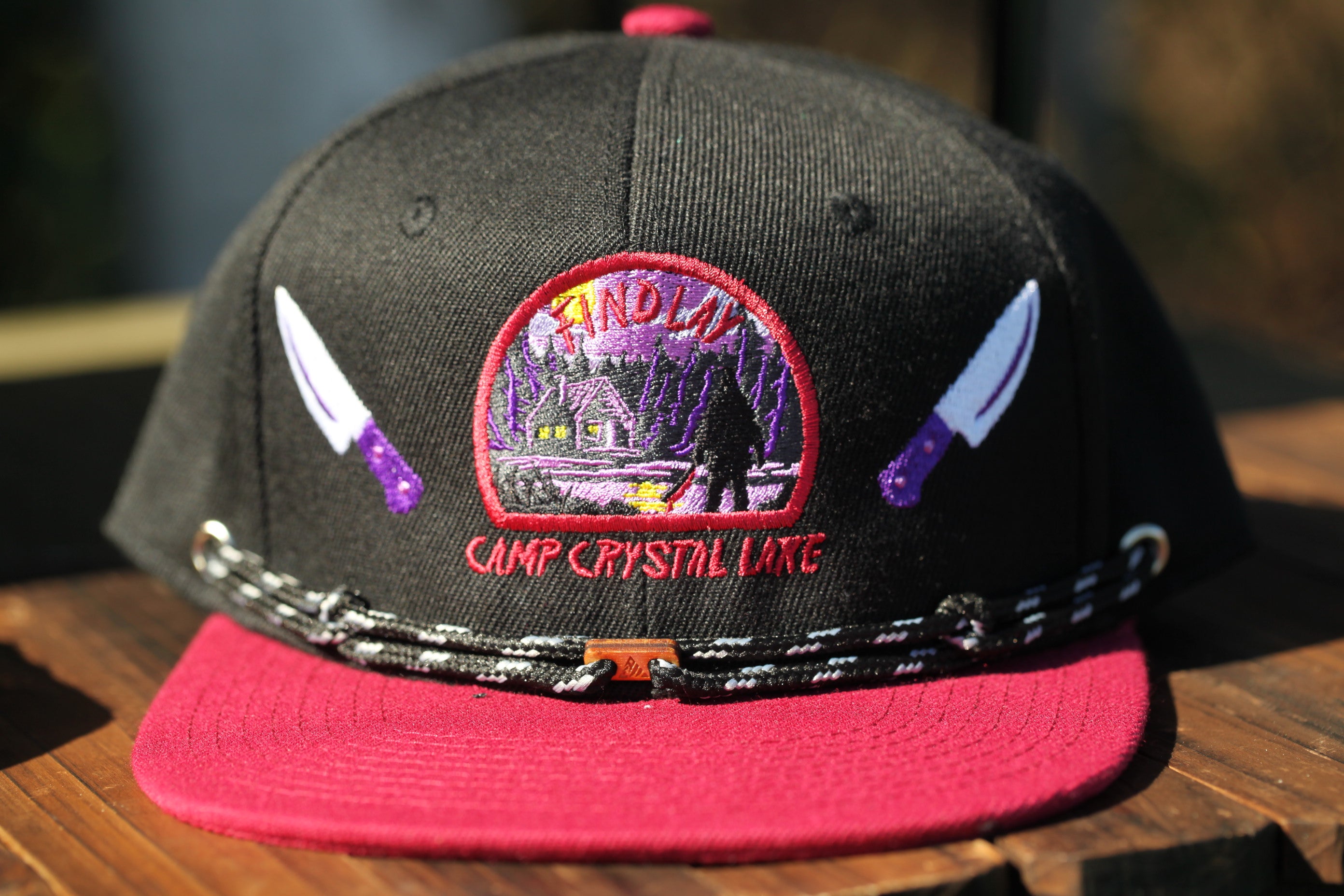 A black baseball cap with a red brim and a patch on the front that says "CAMP CRYSTAL LAKE".