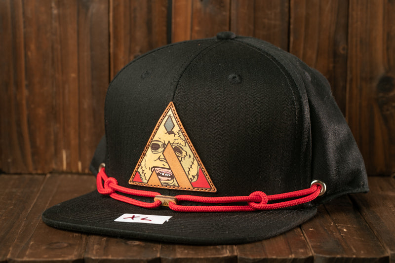 A black baseball cap with a red string and a triangular patch featuring a cartoon character is displayed on a wooden surface.
