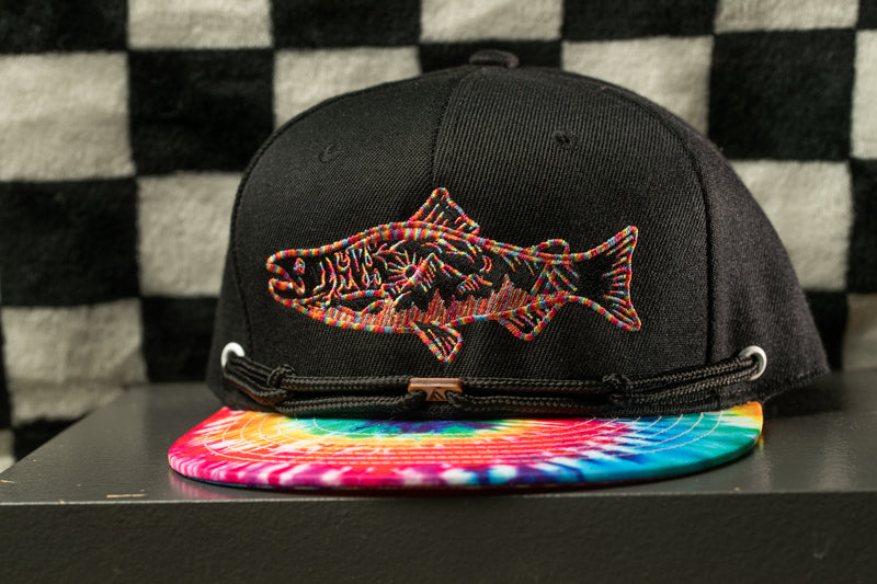 A black baseball cap with a colorful tie-dye pattern and a fish embroidered on the front, placed on a gray surface against a black and white checkered background.