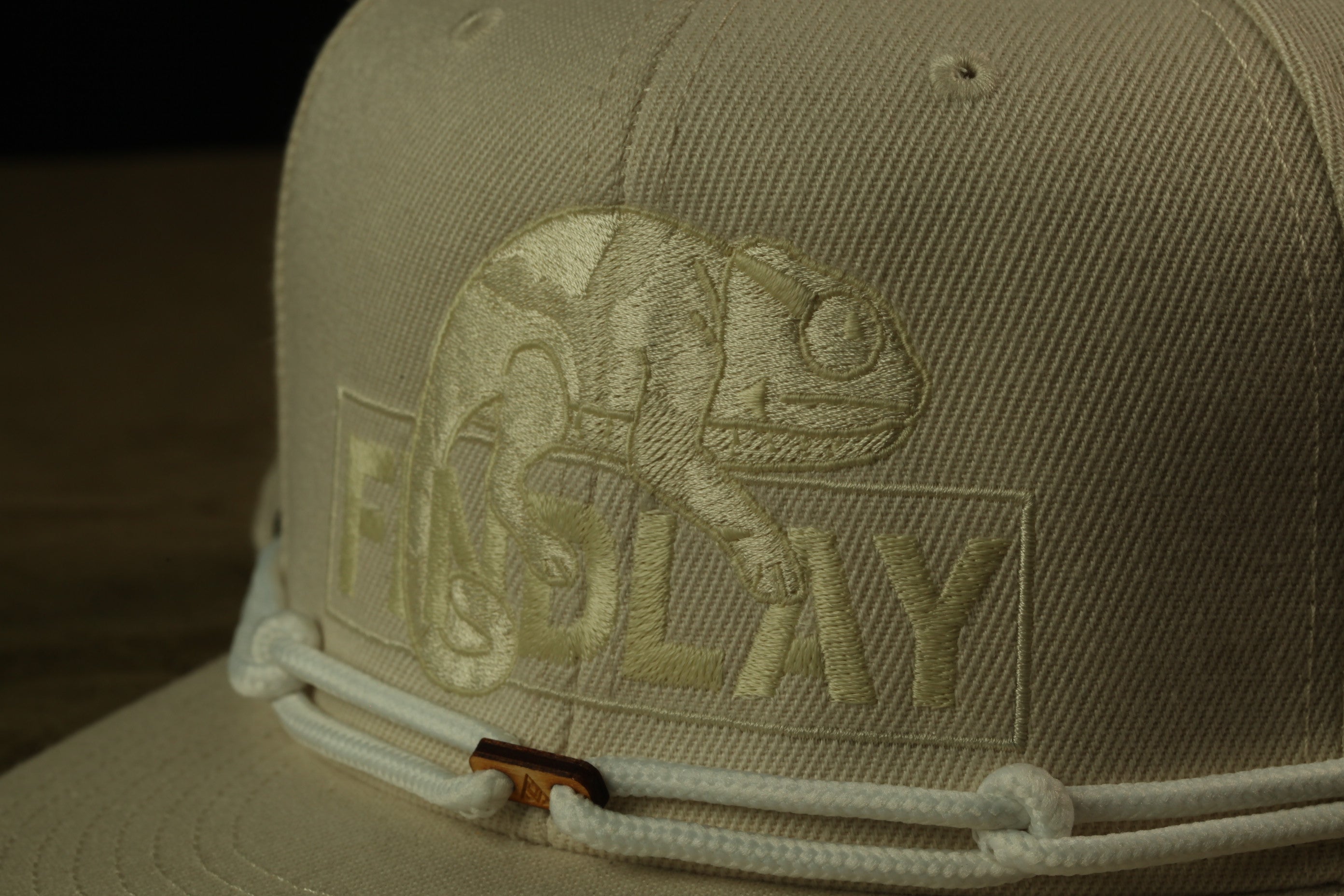 A close-up view of a beige baseball cap with a white embroidered logo on the front.