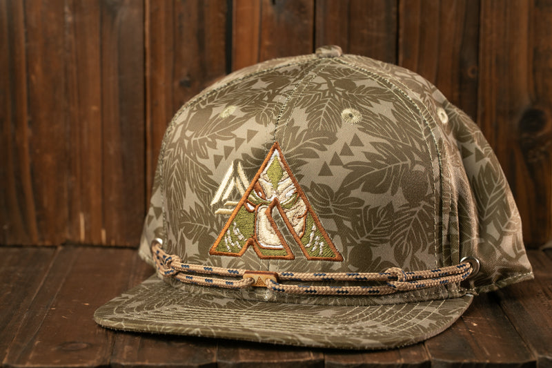 A camouflage-patterned baseball cap with a leaf-like design and a small triangle emblem on the front, resting on a wooden surface.