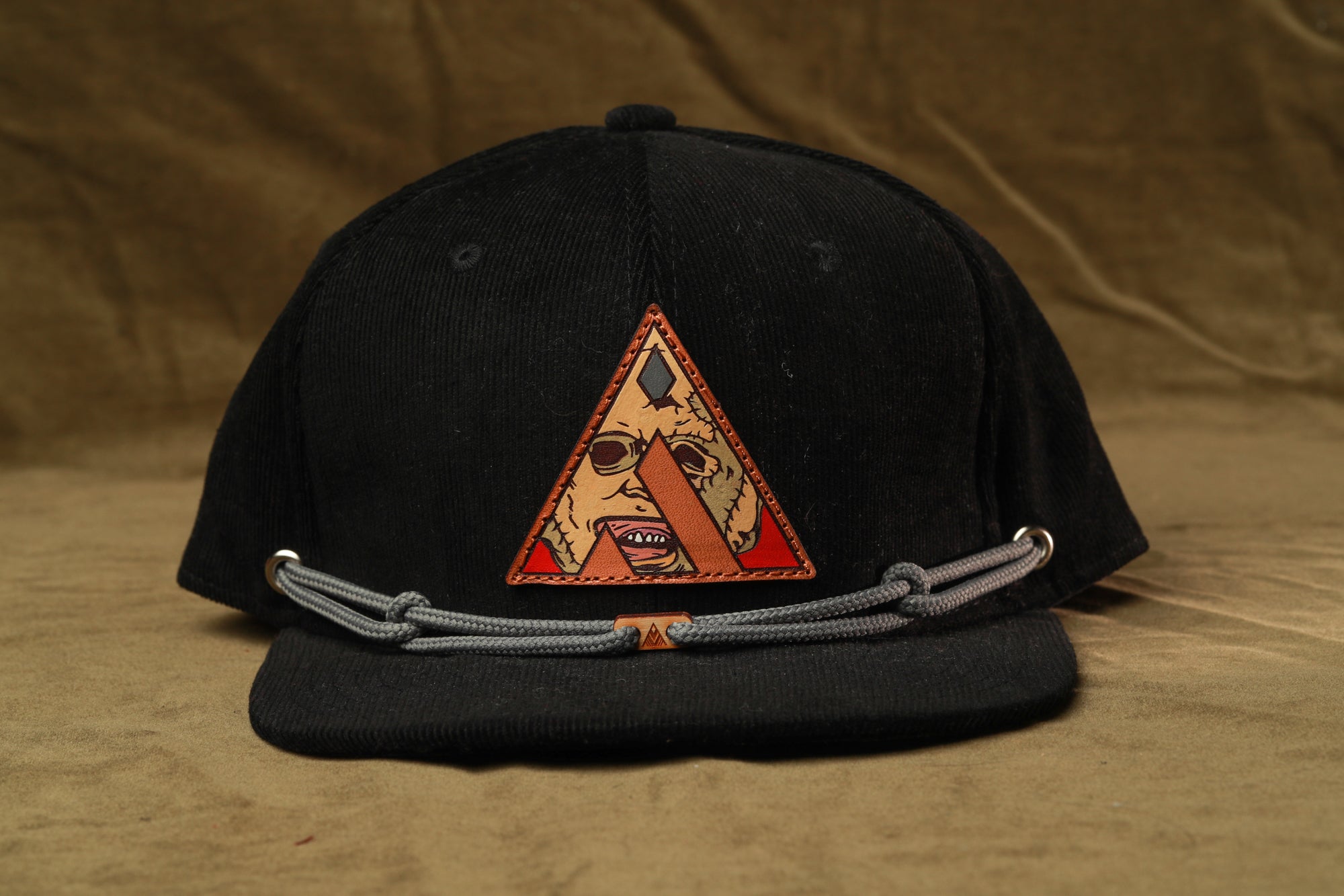 A black baseball cap with a triangular patch featuring a cartoon character on it, and a gray chain strap.