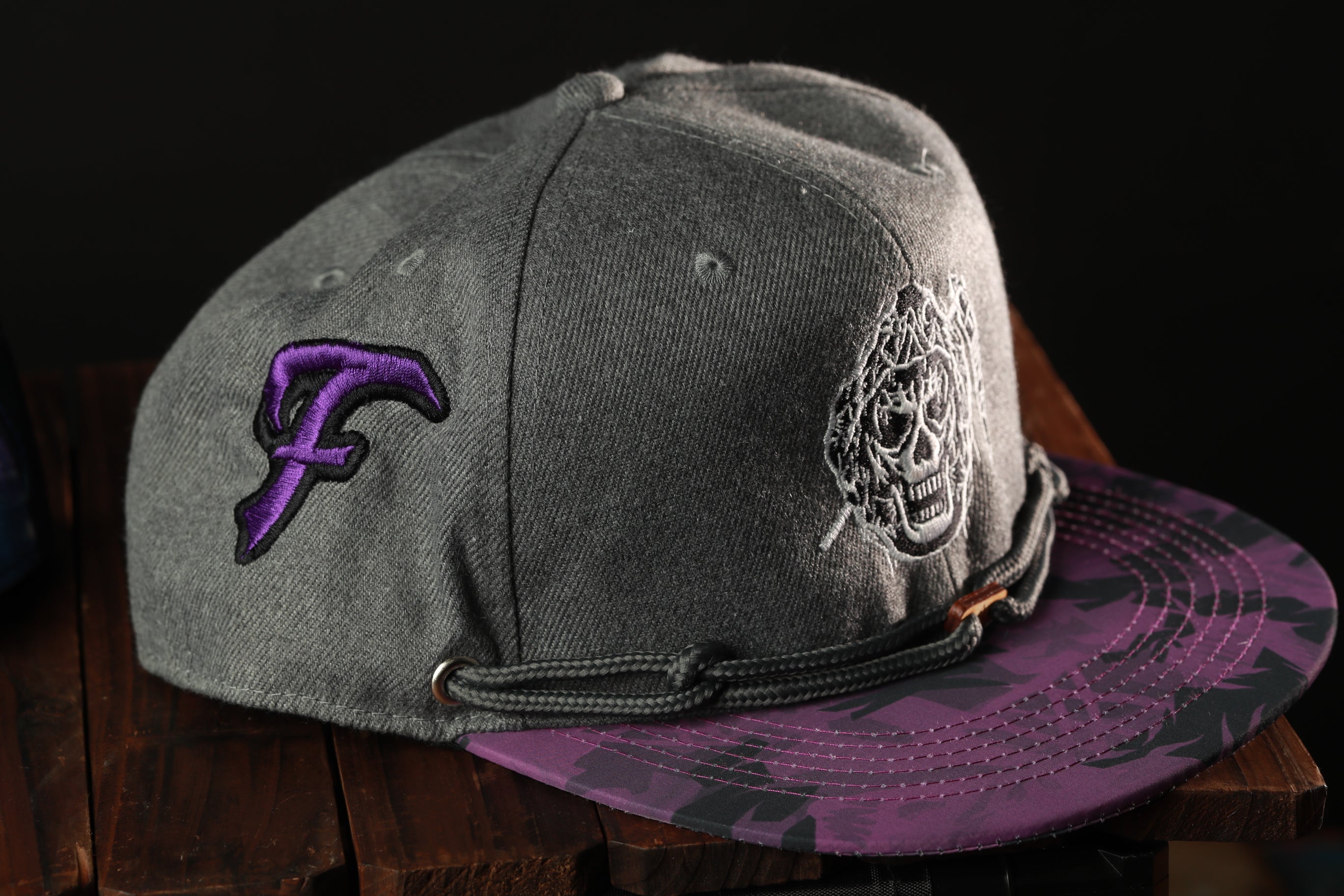 A gray baseball cap with a purple and black camouflage pattern, featuring a skull and crossbones design on the front.