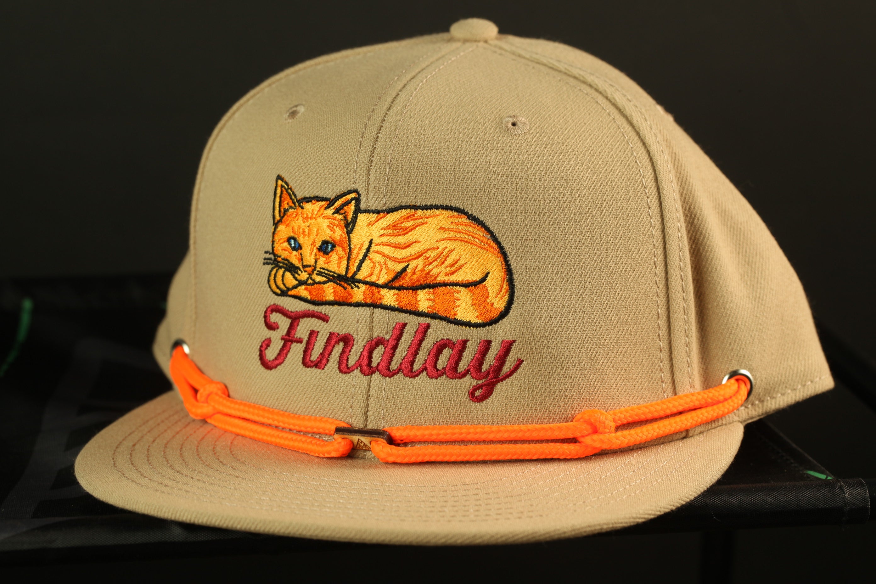 A beige baseball cap with an embroidered image of a cat and the word "Findlay" printed on it.