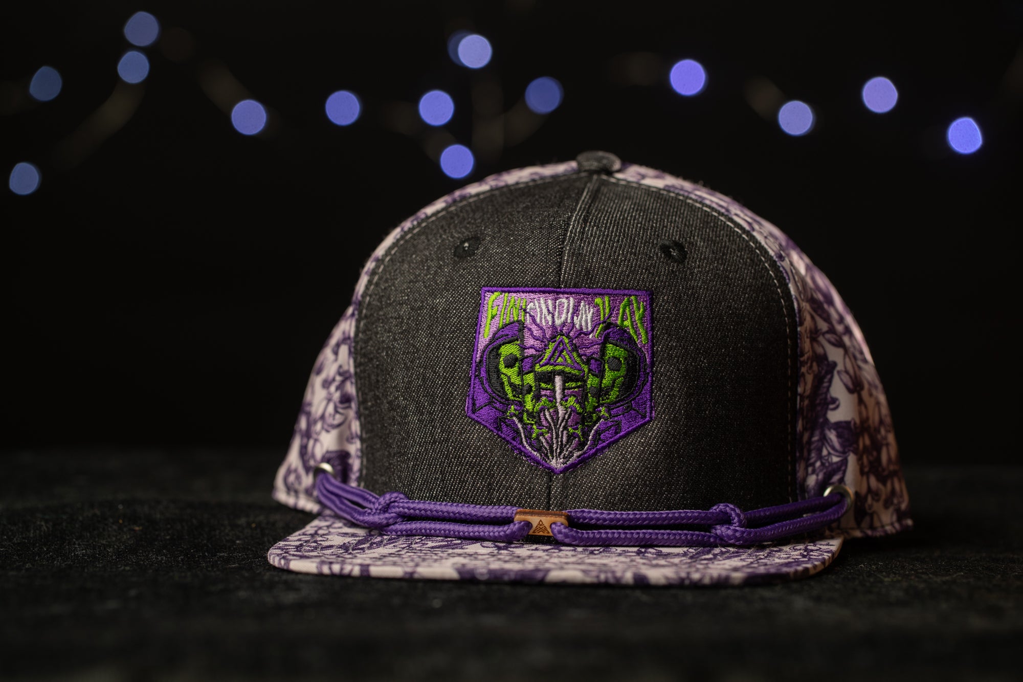A black baseball cap with a purple and white floral pattern, featuring a purple patch with a skull and crossbones design.