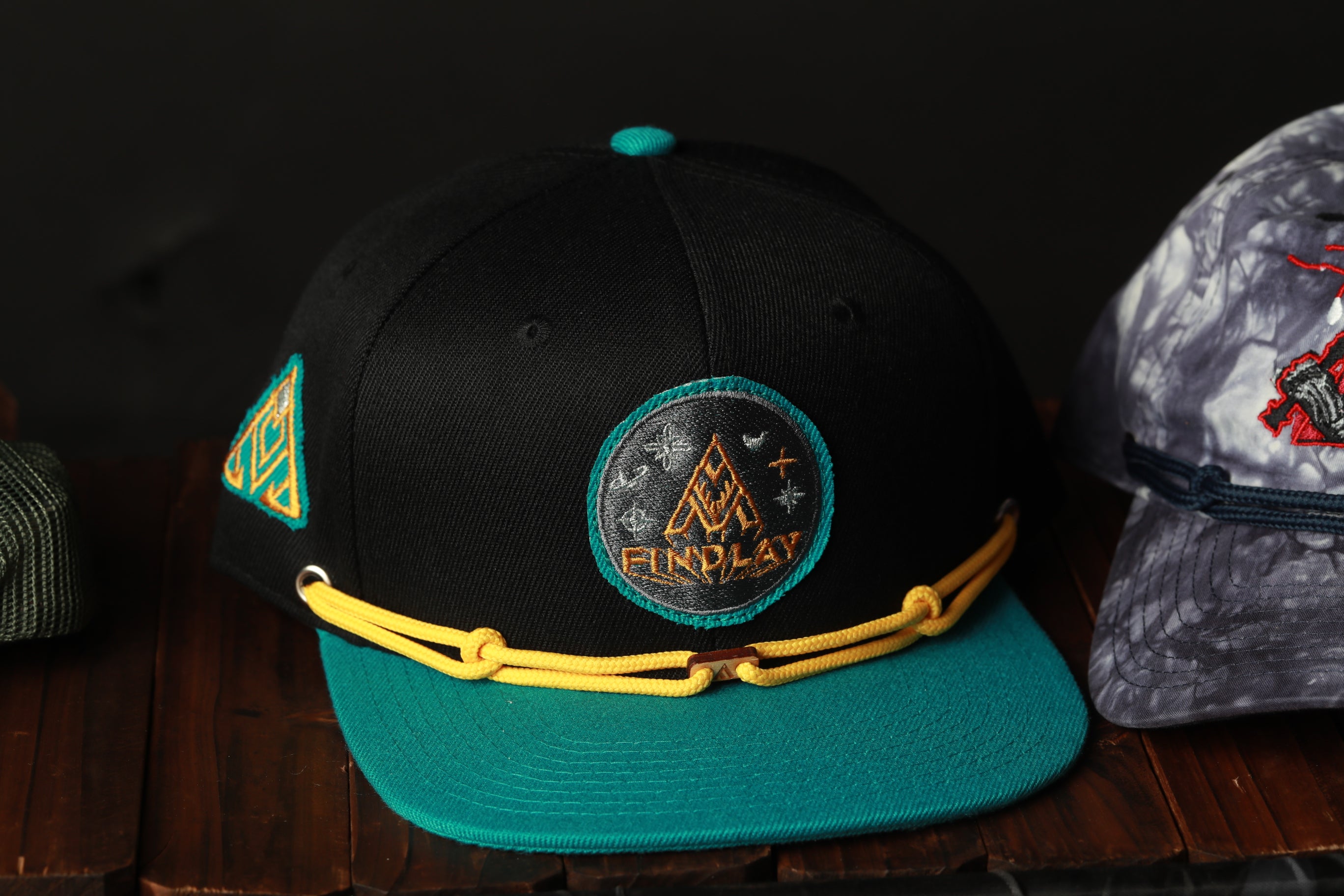 A black baseball cap with a teal brim and yellow straps, featuring a circular patch with the word "FIND" and a mountain-like logo.