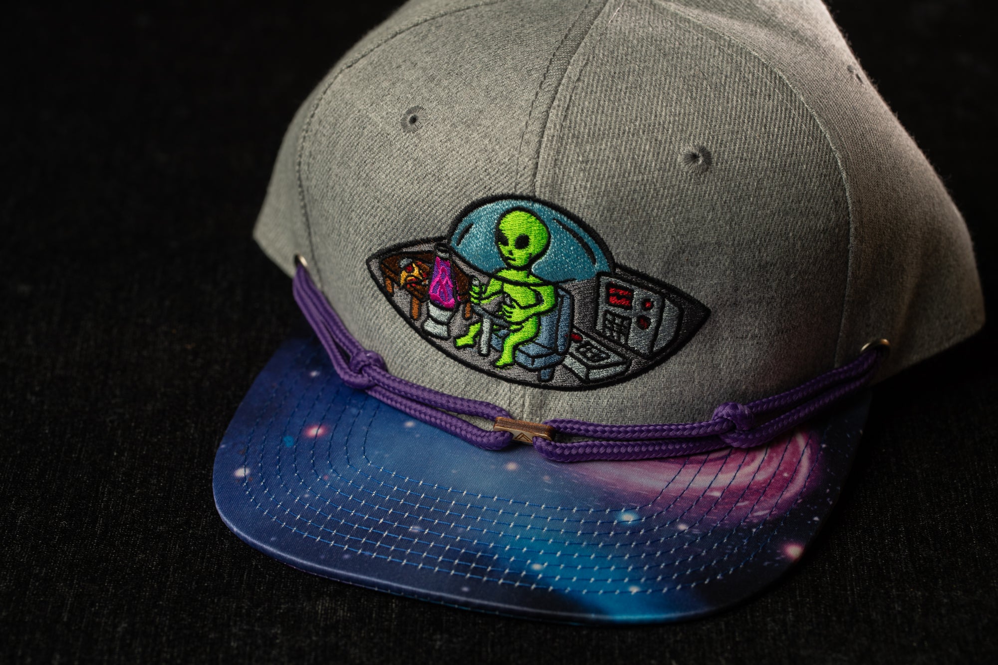 A gray baseball cap with a purple brim and a colorful alien embroidered on the front.