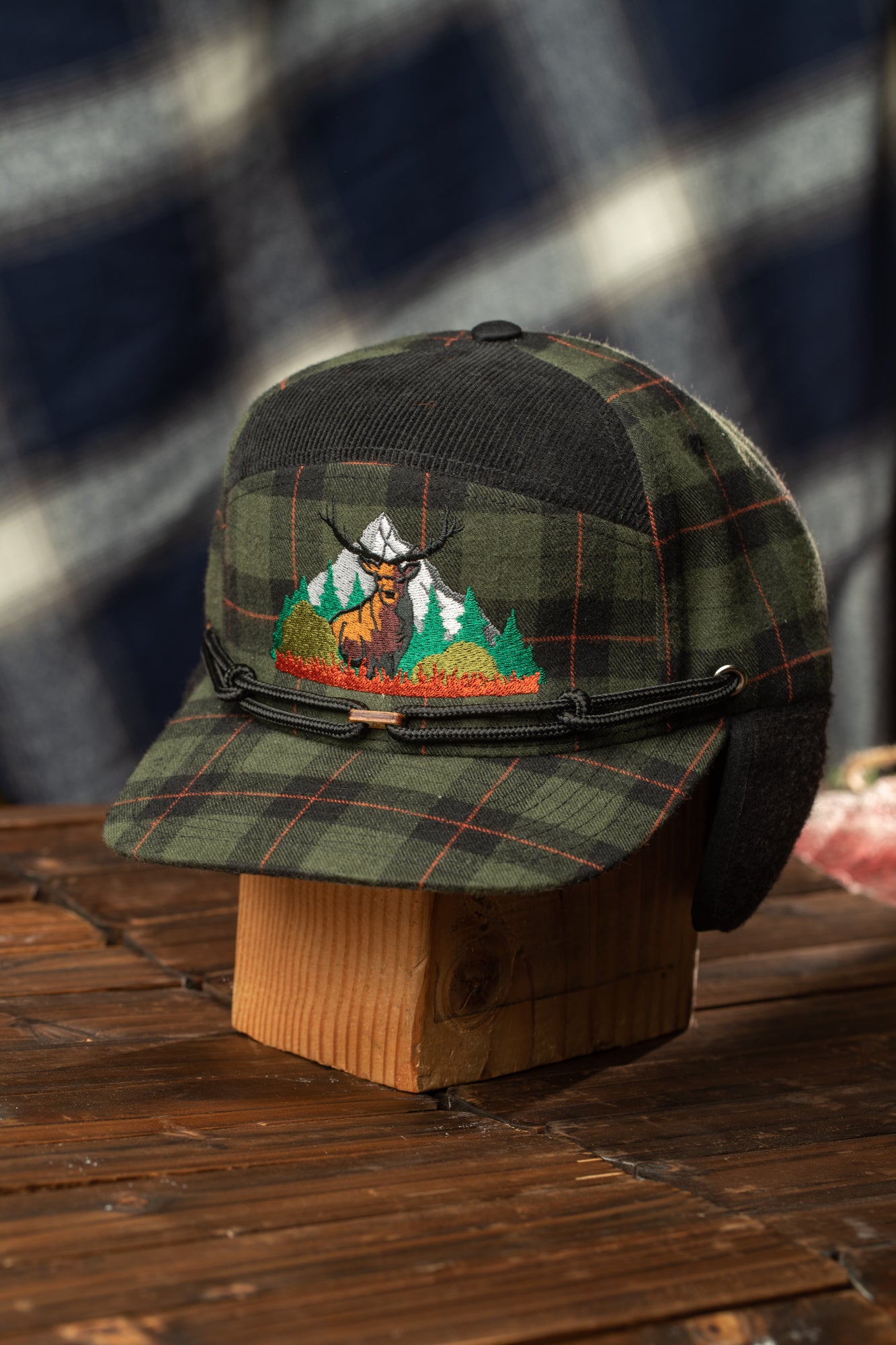 A plaid hat with a mountain landscape embroidered on it, placed on a wooden stand.