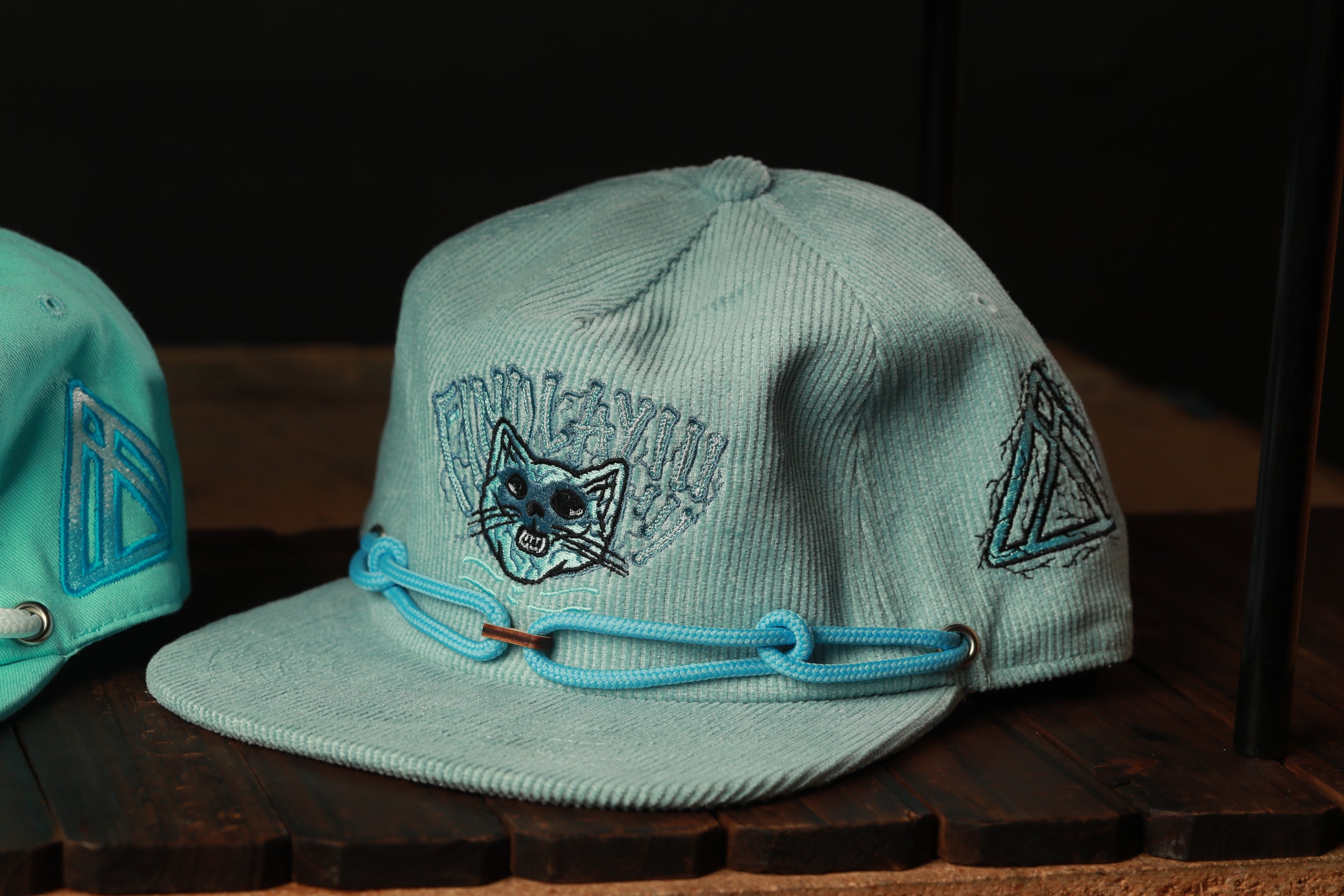A light blue baseball cap with a cat design and a blue strap is displayed on a wooden surface.