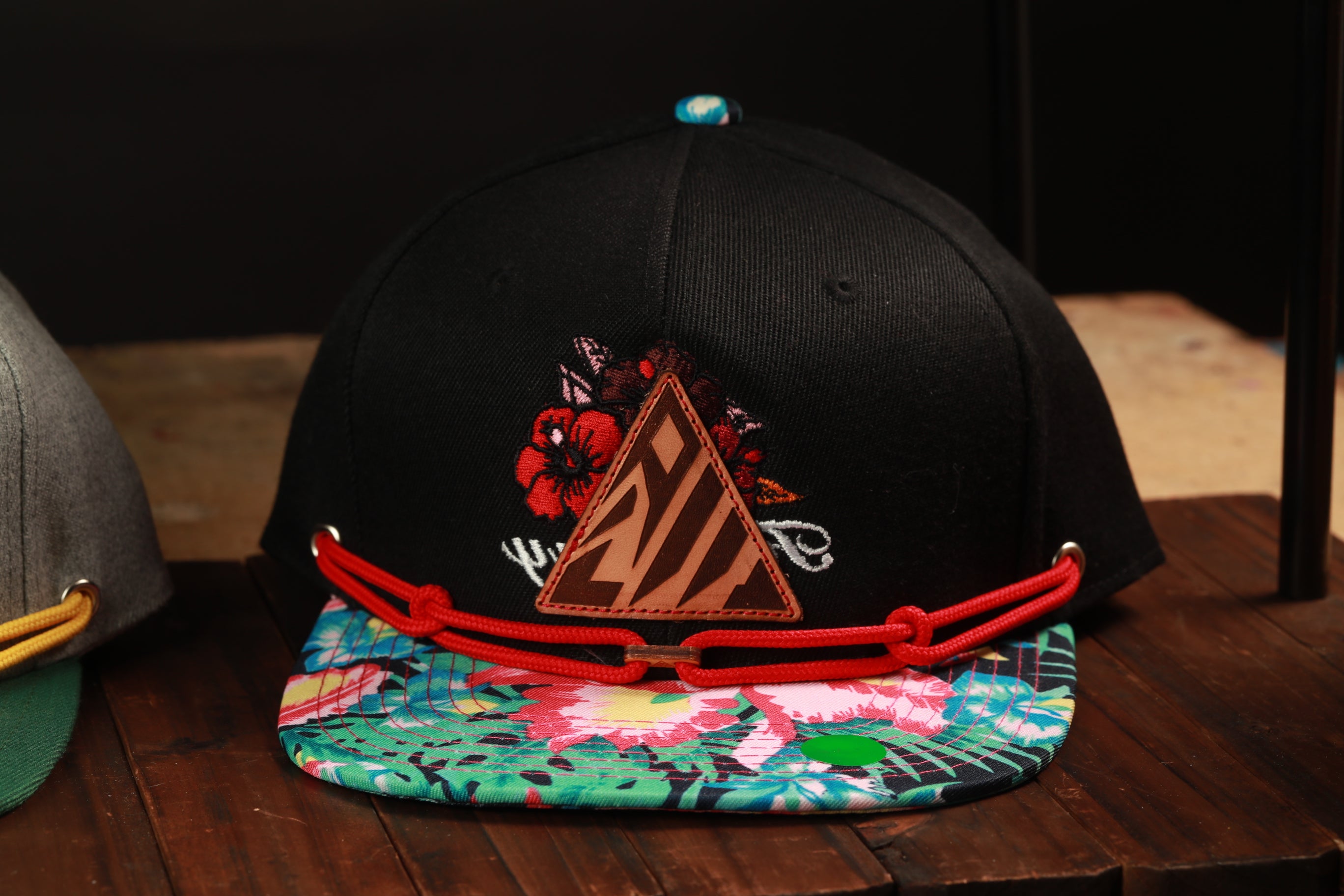 A black baseball cap with a floral pattern and a red strap is displayed on a wooden surface.