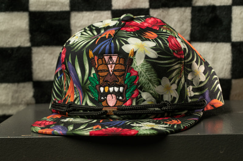 A colorful, patterned baseball cap with a tropical design, including flowers and leaves, is displayed against a checkered background.