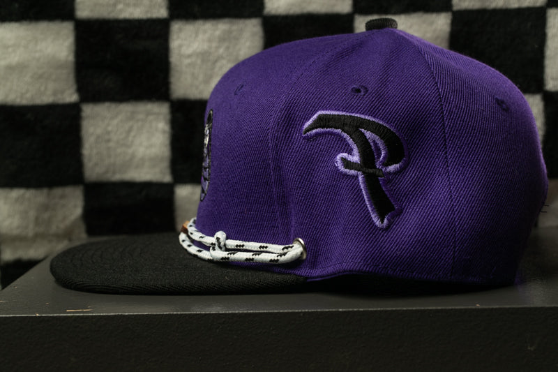 A purple baseball cap with a black logo on the front, placed on a black surface against a checkered background.
