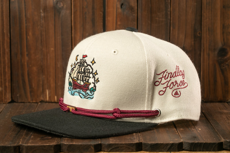 A white baseball cap with a black brim and a maroon band, featuring an embroidered design of a ship on the front.