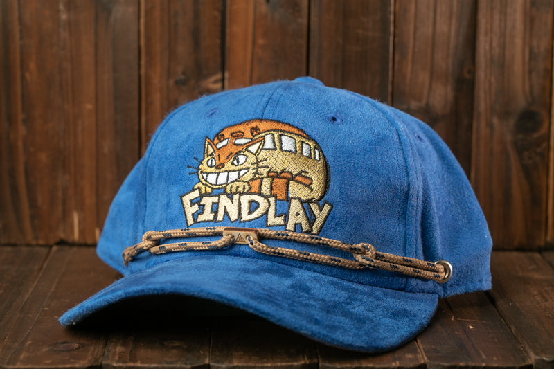 A blue baseball cap with a cat design and the word "FINDlay" printed on it, sitting on a wooden surface.