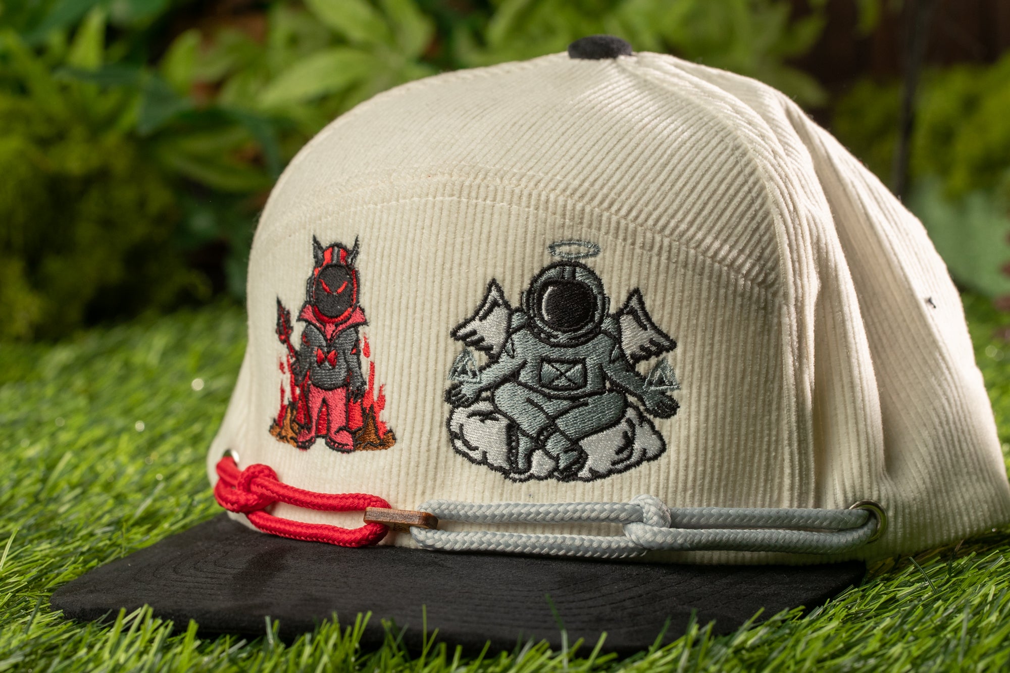 A beige baseball cap with two embroidered characters on it, one of a devil and one of an angel, sitting on a grassy surface.