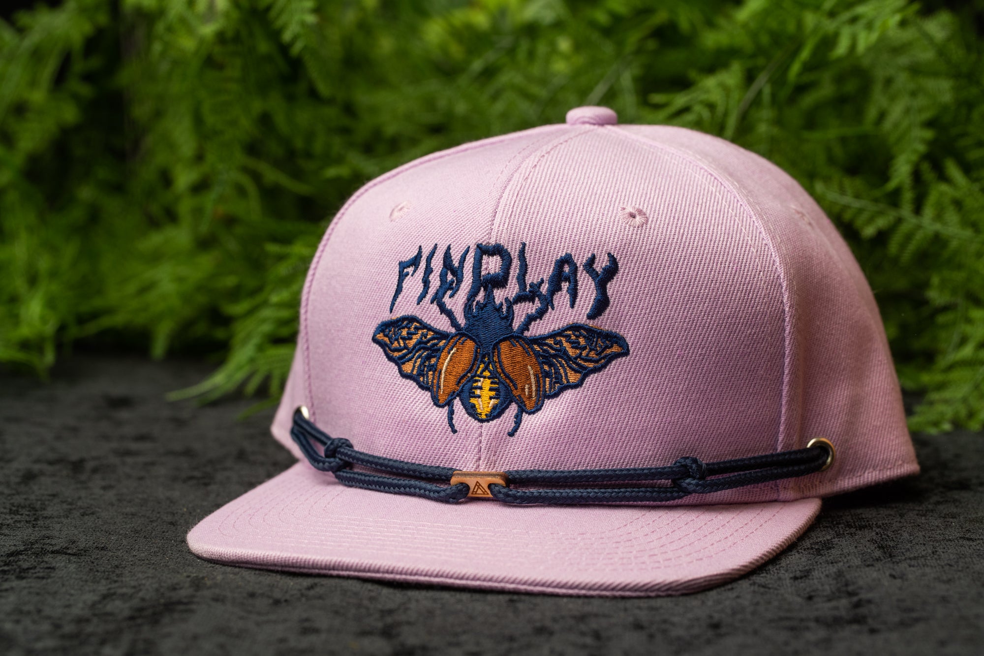 A pink baseball cap with a graphic of a bee and the text "FINALLY" printed on it, placed on a surface with green foliage in the background.
