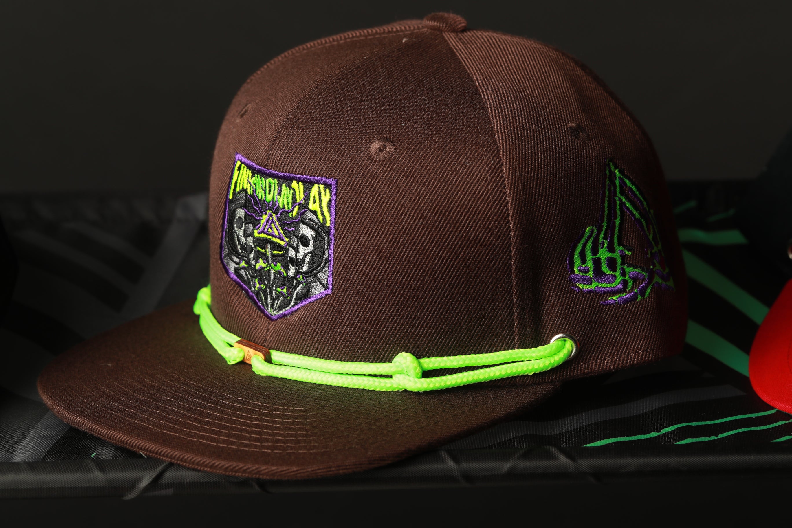 A brown baseball cap with a green neon string and a purple and green patch with a skull and crossbones design on the front.