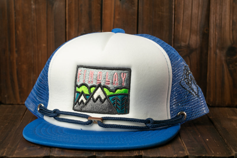 A blue and white baseball cap with a mountain landscape embroidered on it, placed on a wooden surface.