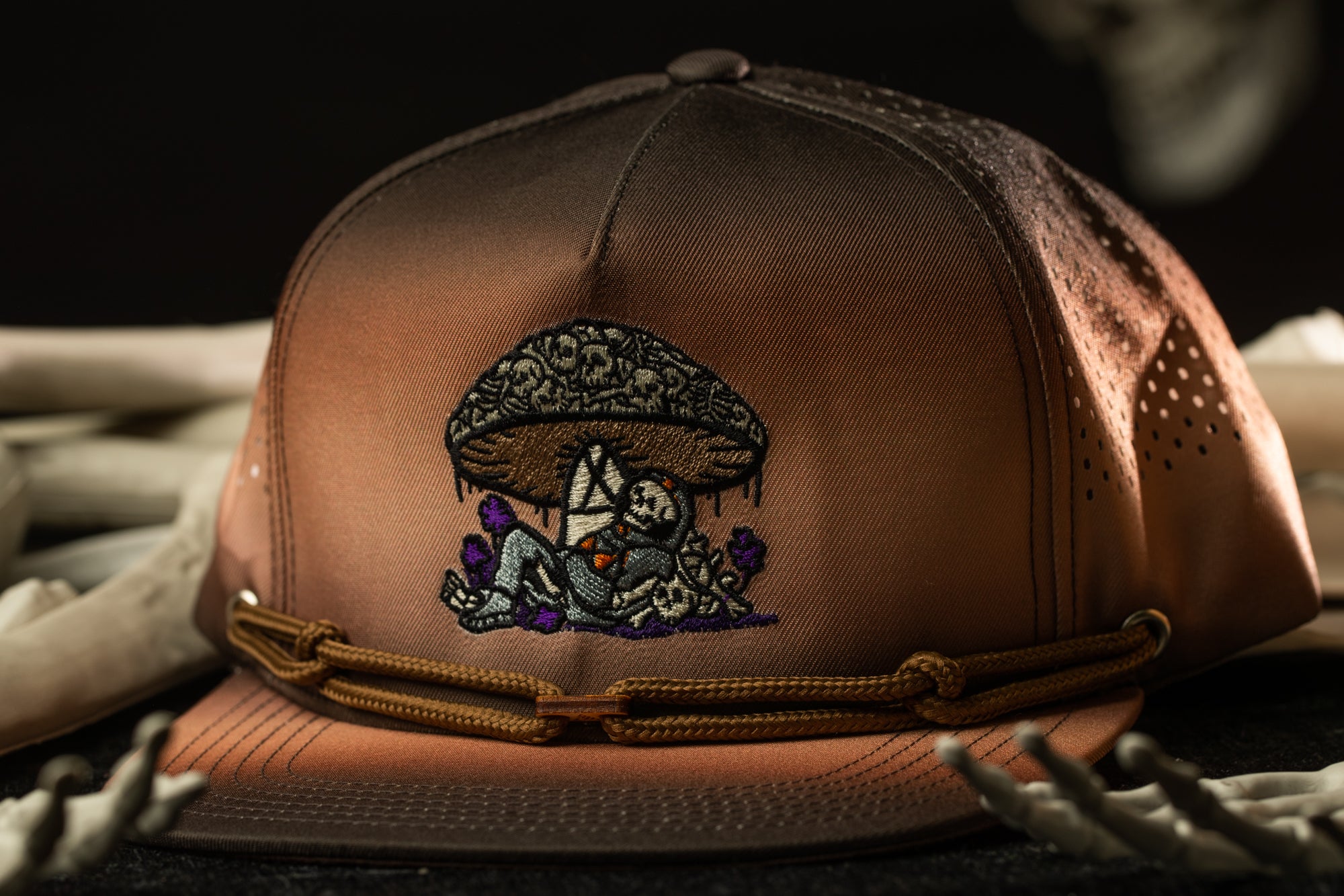 A brown baseball cap with an embroidered design on the front, featuring a mushroom and a person.