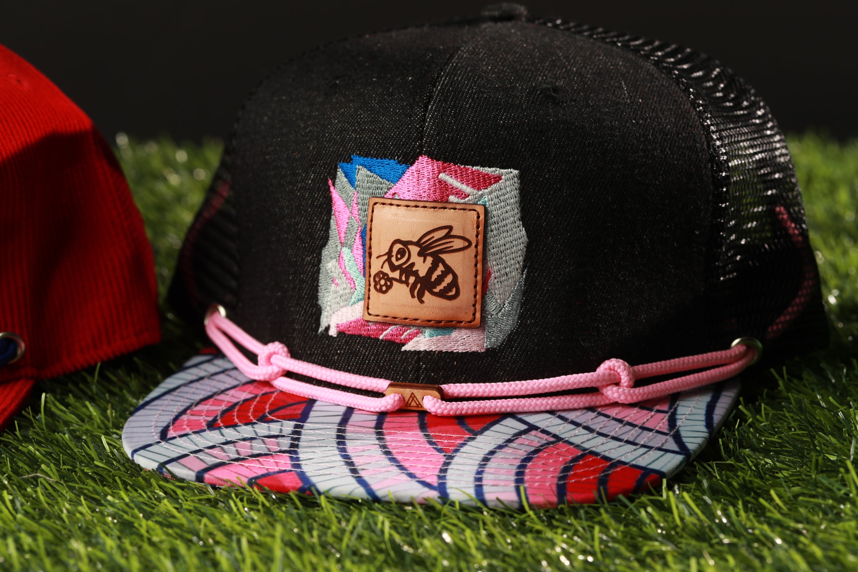 A black baseball cap with a colorful pattern and a small embroidered patch featuring a bee design, placed on a grassy surface.