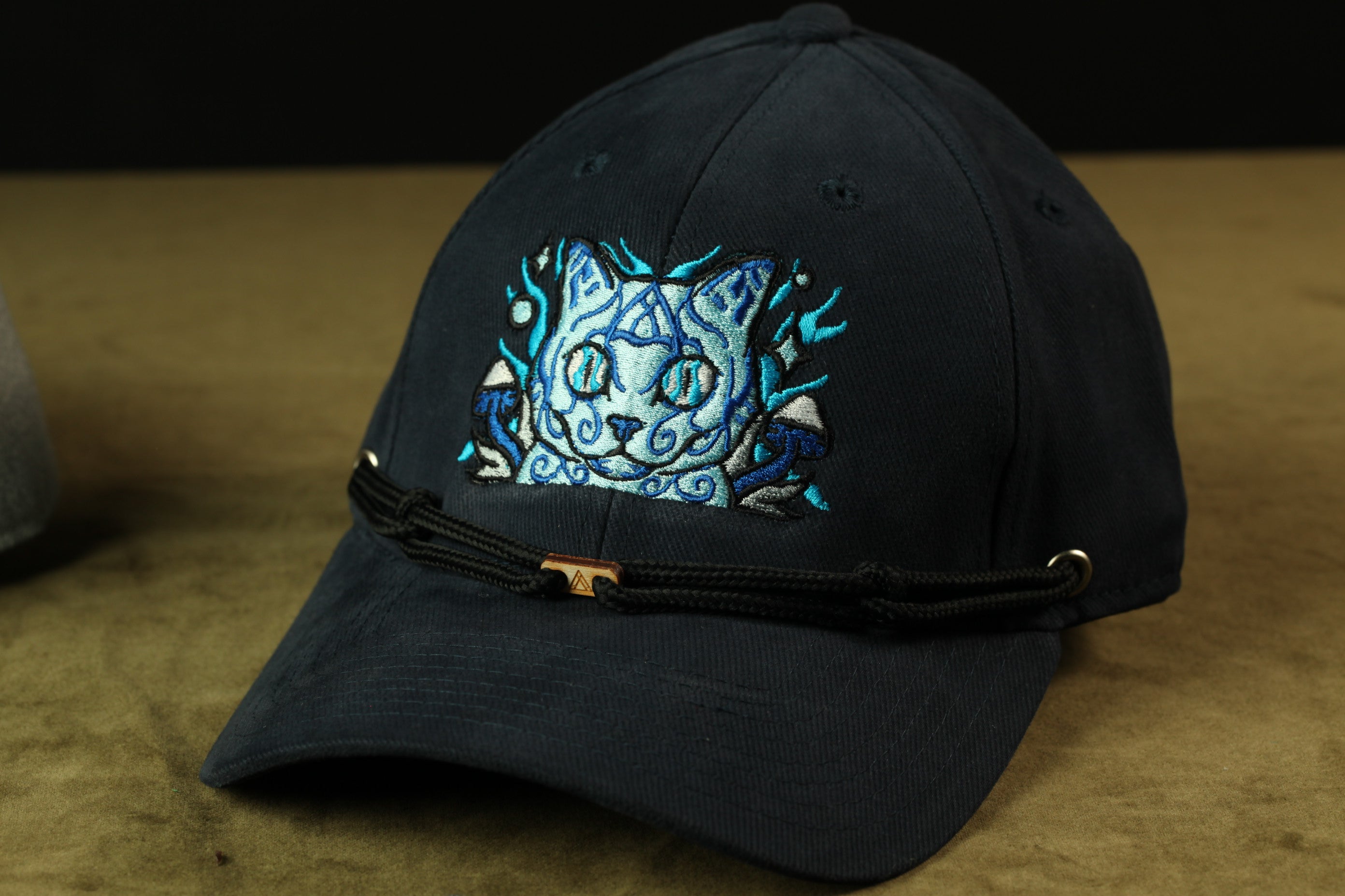 A black baseball cap with a blue cat embroidered on the front, sitting on a brown surface.