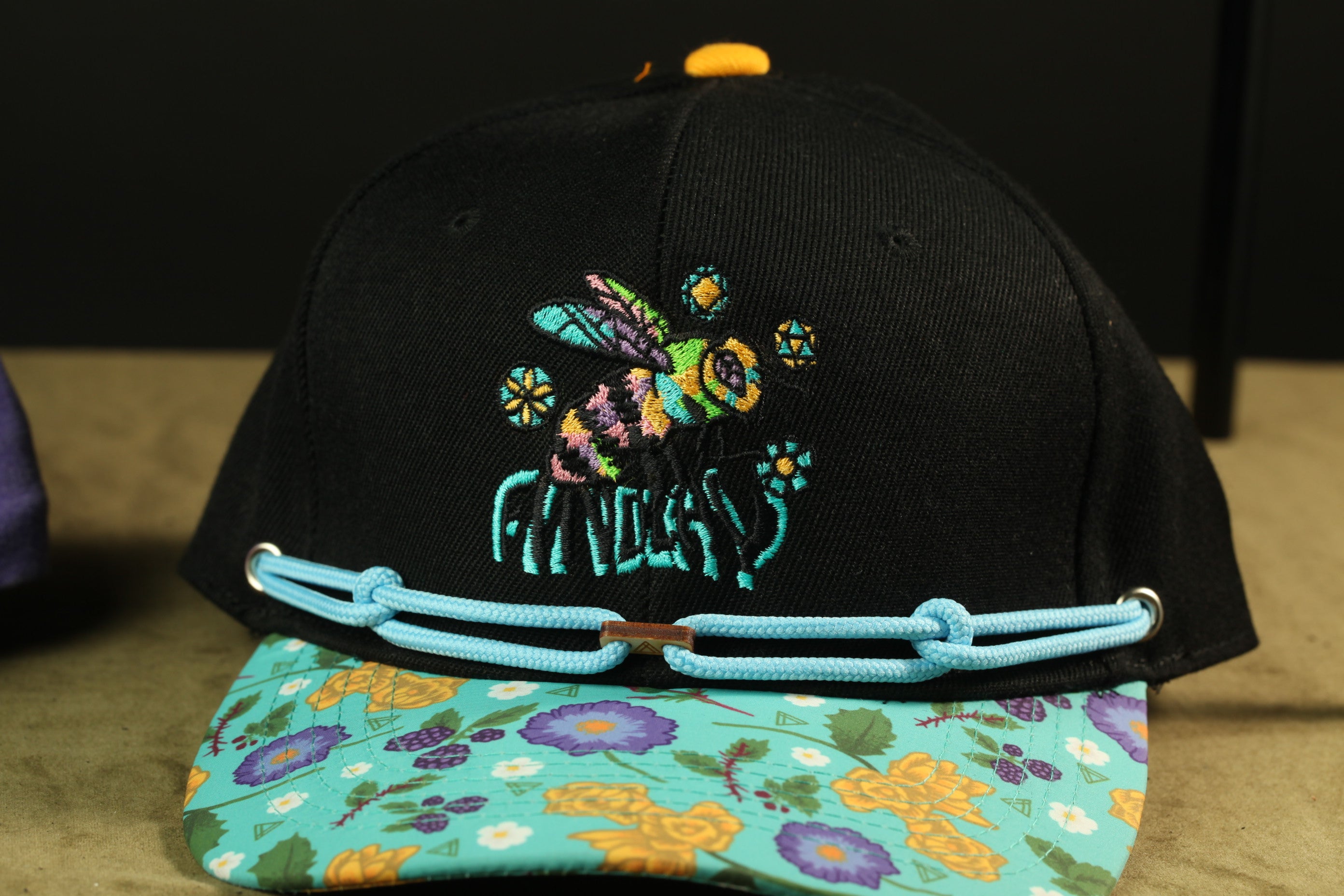 A black baseball cap with a colorful floral pattern and a bee embroidered on it, along with a blue lace trim.