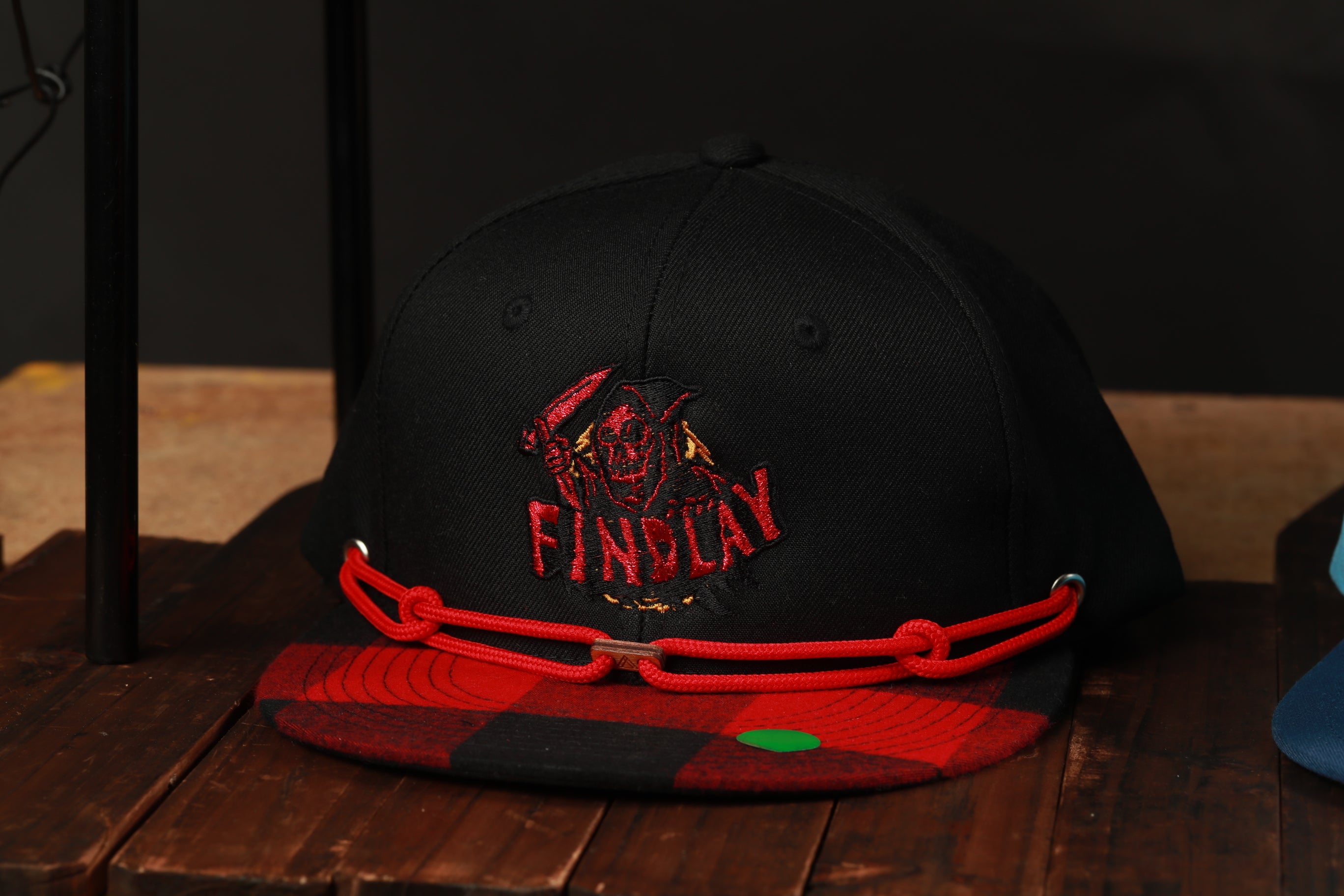 A black baseball cap with a red plaid pattern and a red string around the brim, placed on a wooden surface.