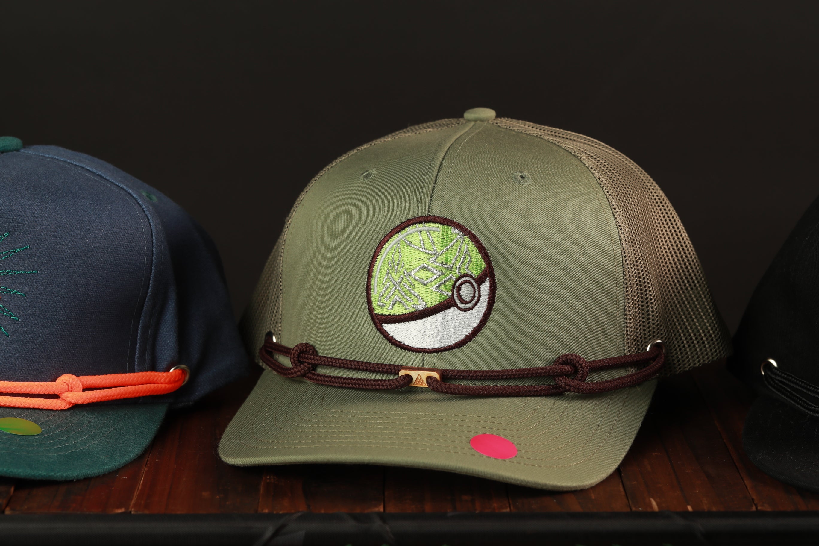 A green baseball cap with a Pokemon patch on it, placed on a wooden surface next to other hats.