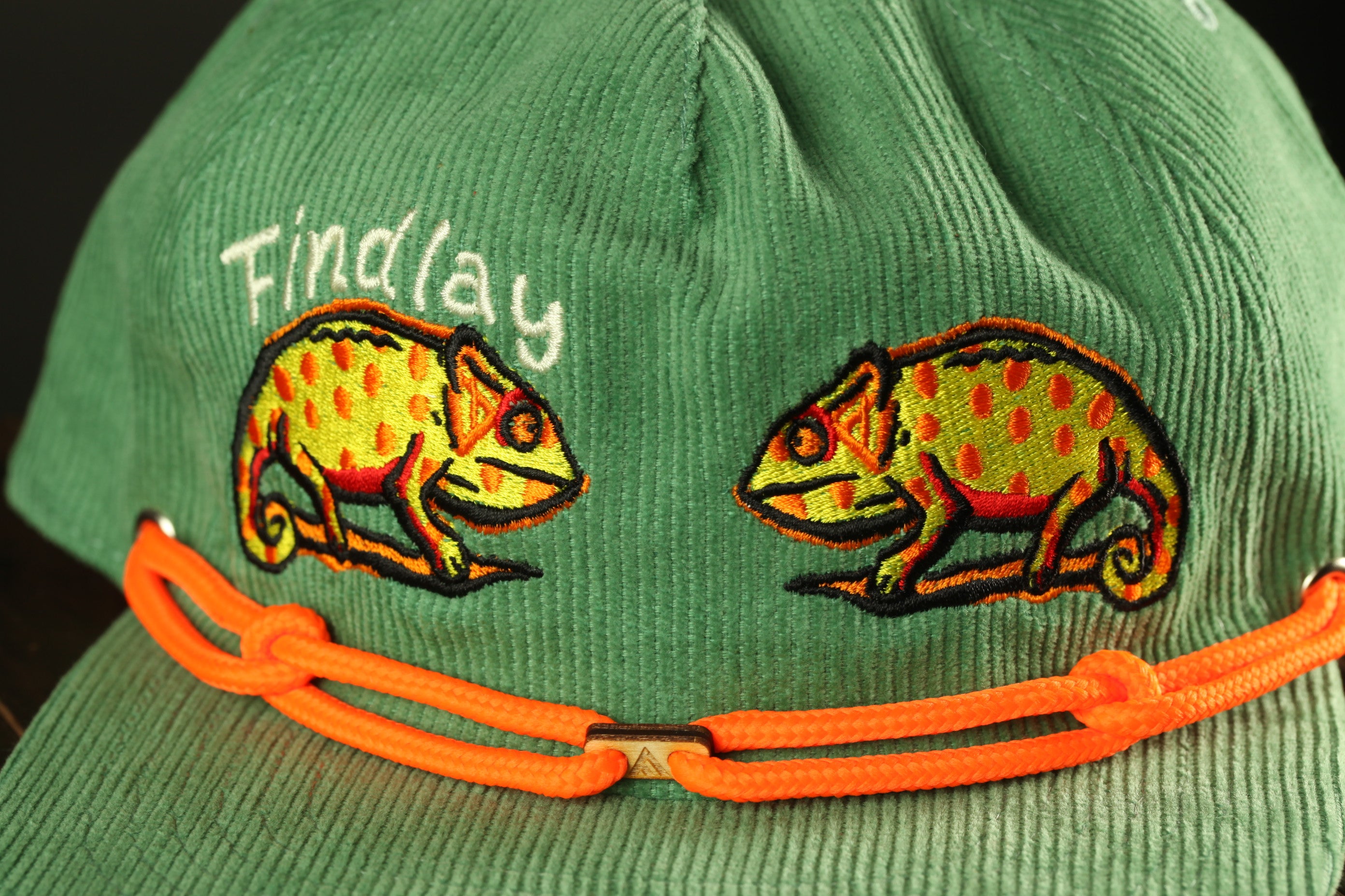 A green baseball cap with orange braided straps and two chameleon designs on the front.