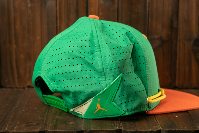 A green and orange baseball cap with a perforated pattern and a small orange logo on the front, resting on a wooden surface.