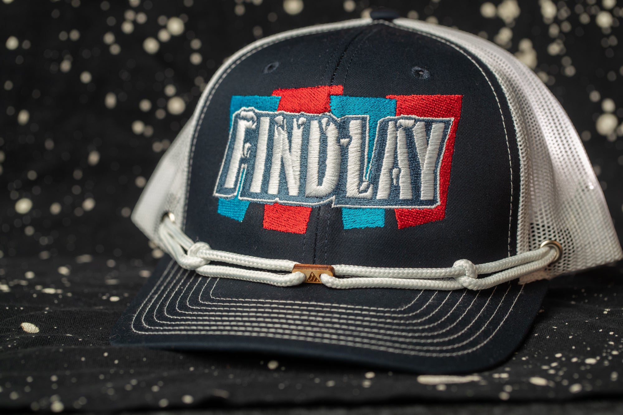 A black baseball cap with the word "FINDlay" embroidered on it, set against a black background with white speckles.