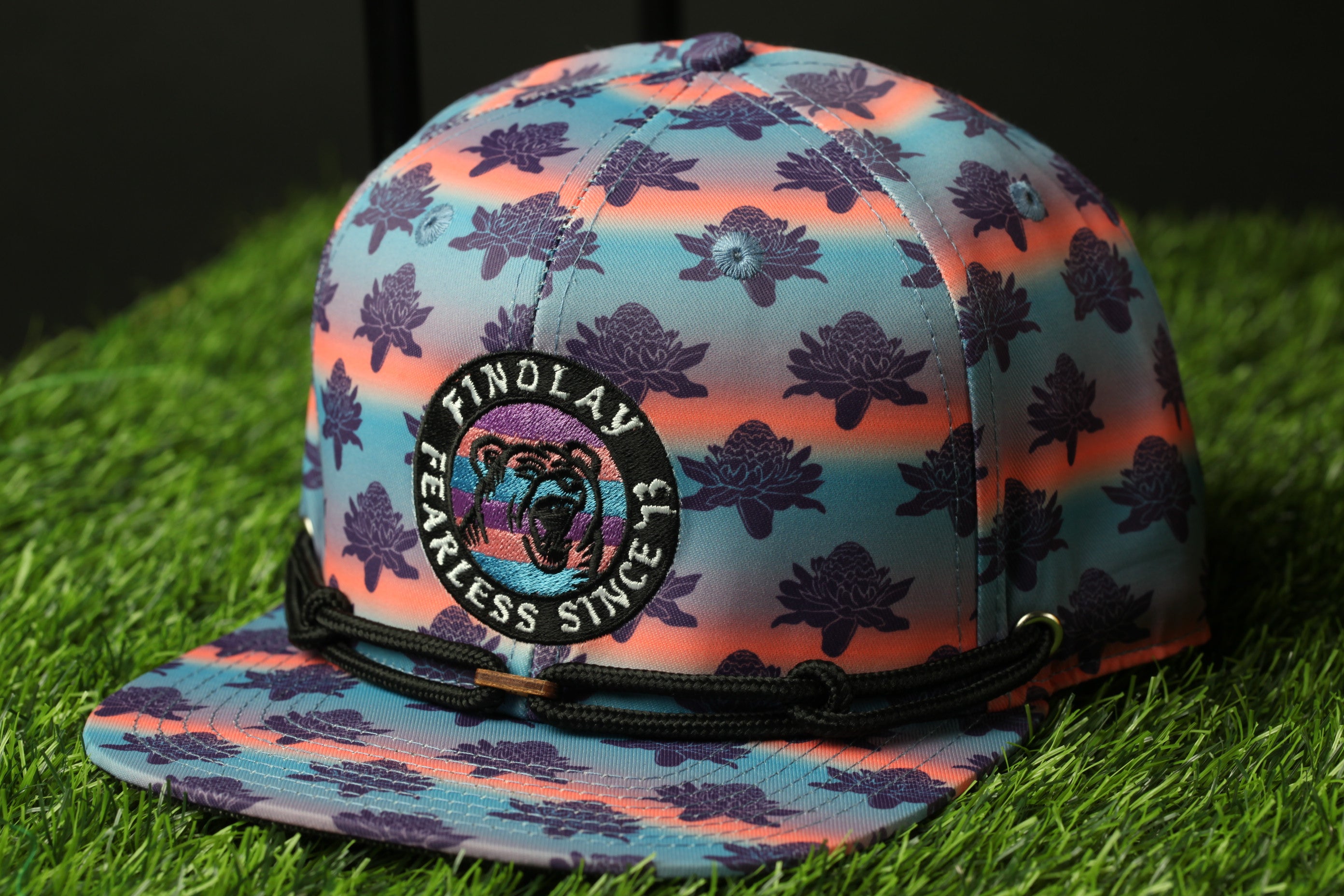 A colorful baseball cap with a pattern of purple and orange flowers, featuring a circular logo with a bear and the text "FINOLAY FEARLESS SINCE 13".