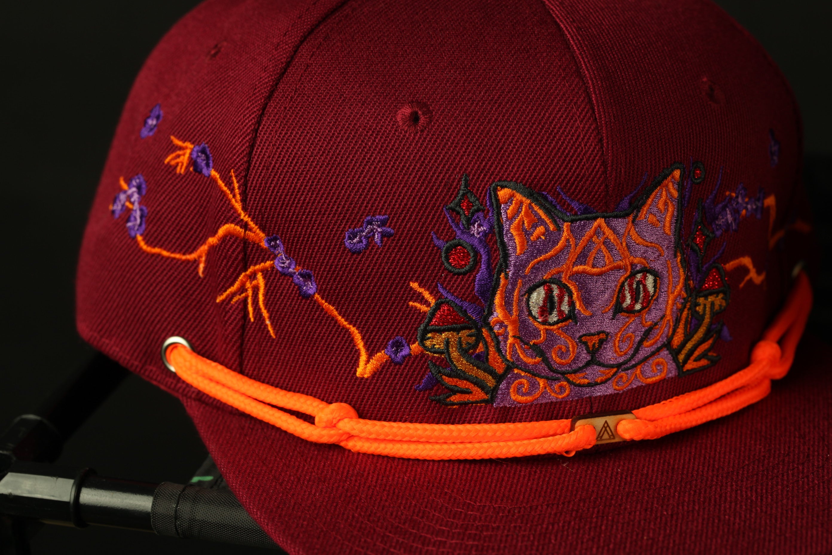 A close-up image of a burgundy baseball cap with a cat embroidered on it, featuring a colorful and intricate design.