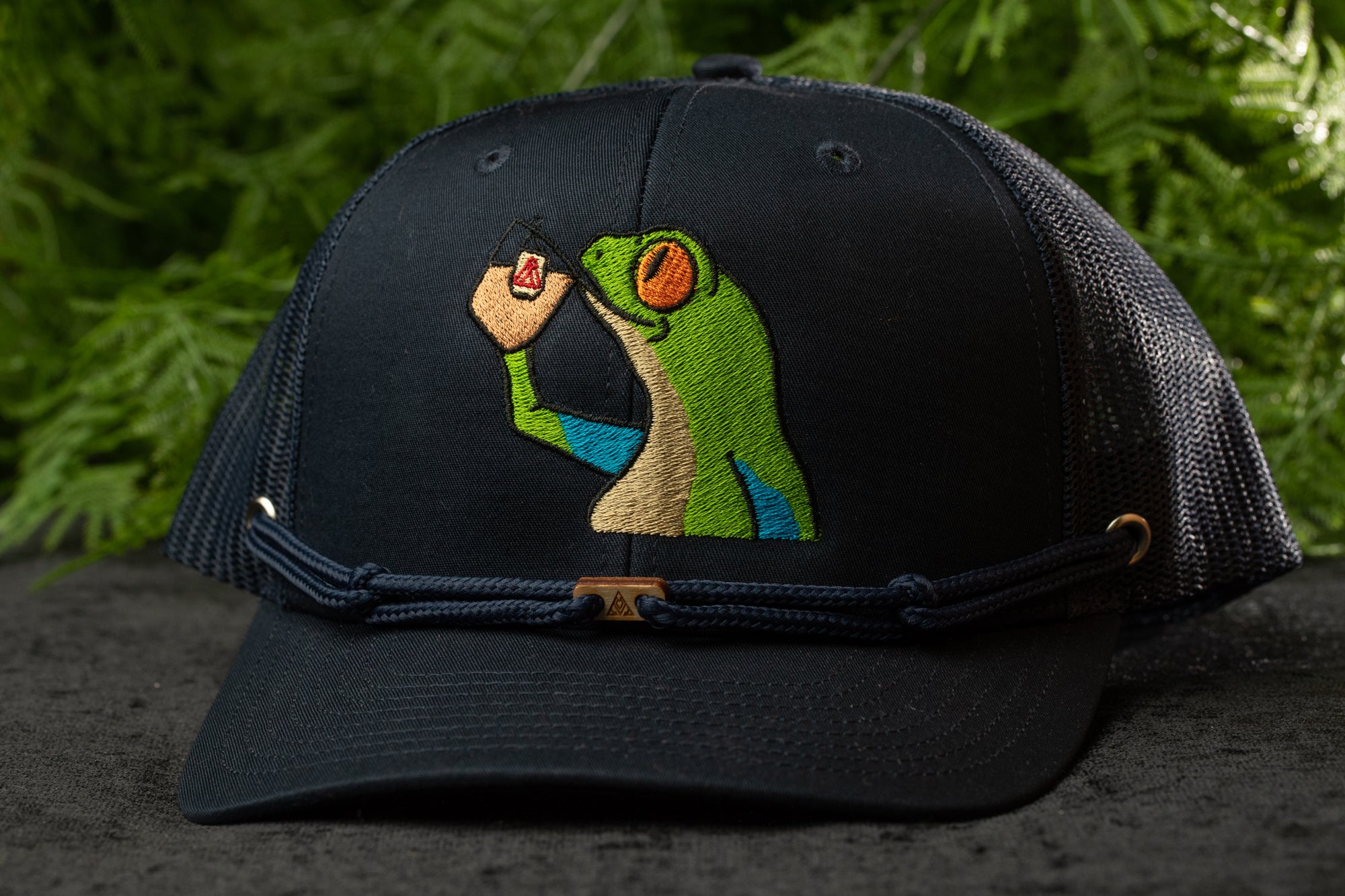 A black baseball cap with a colorful frog embroidered on it, set against a blurred green background.