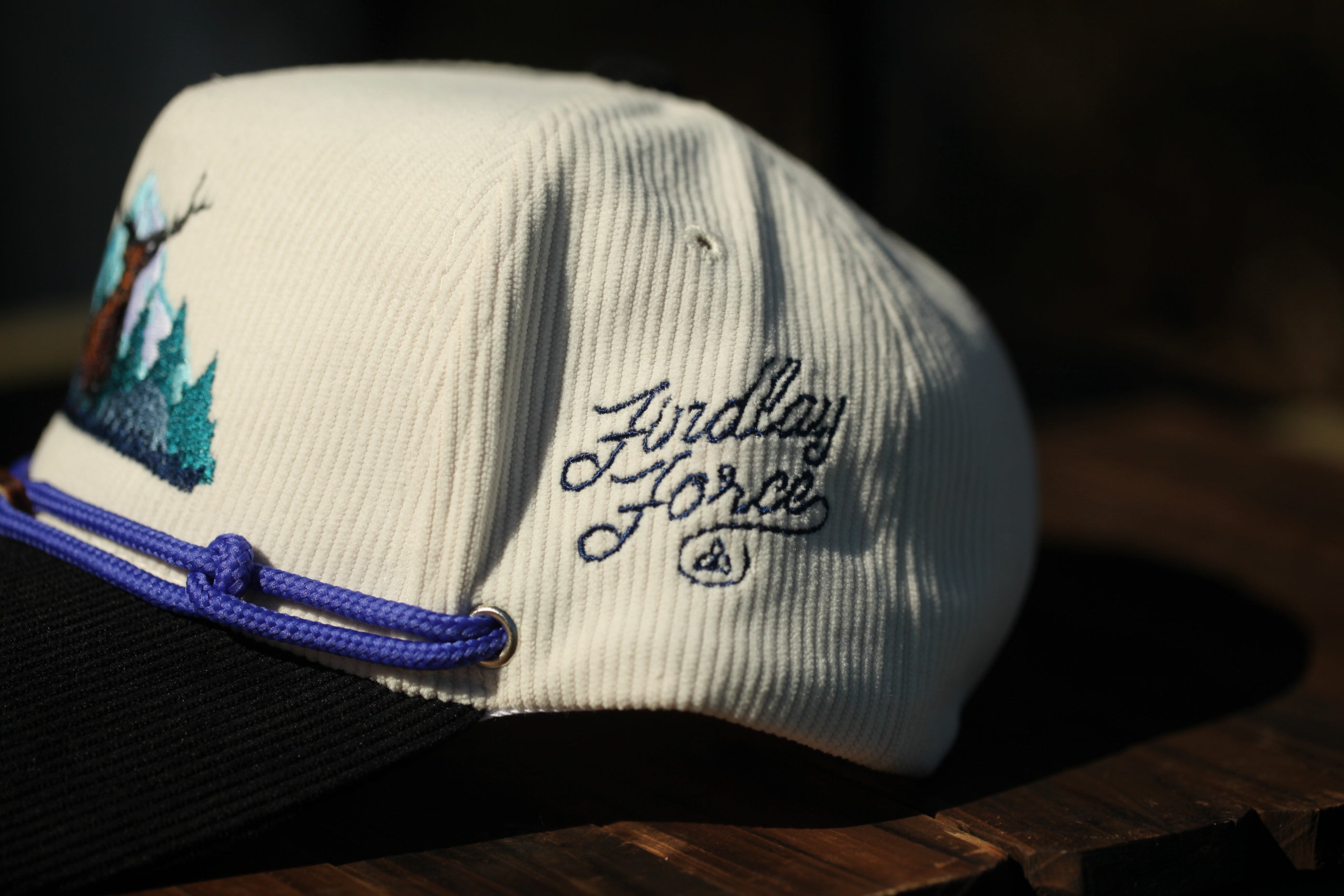 A white baseball cap with blue embroidered text and a graphic design on the front, resting on a wooden surface.