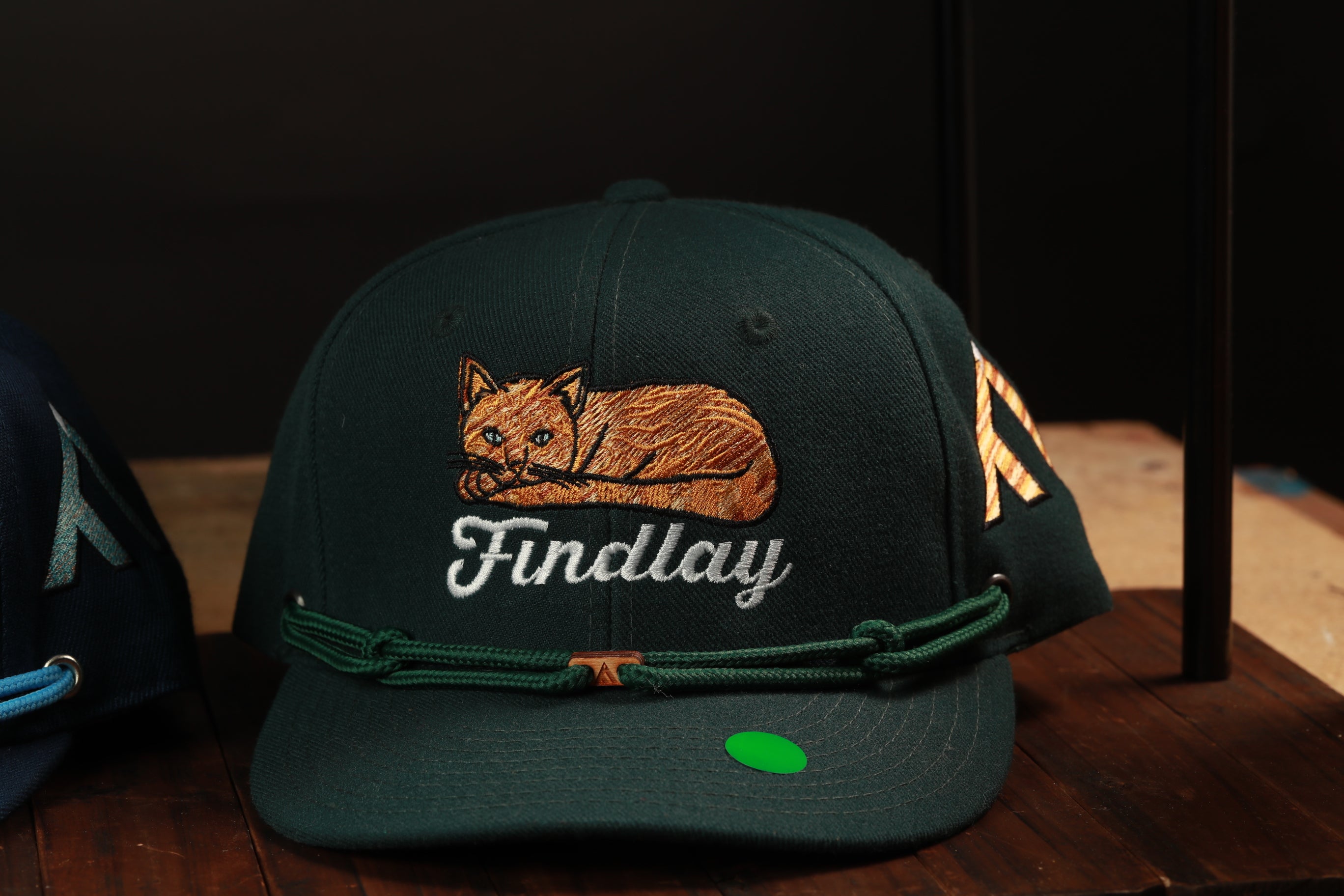 A dark green baseball cap with a cat embroidered on it, sitting on a wooden surface.