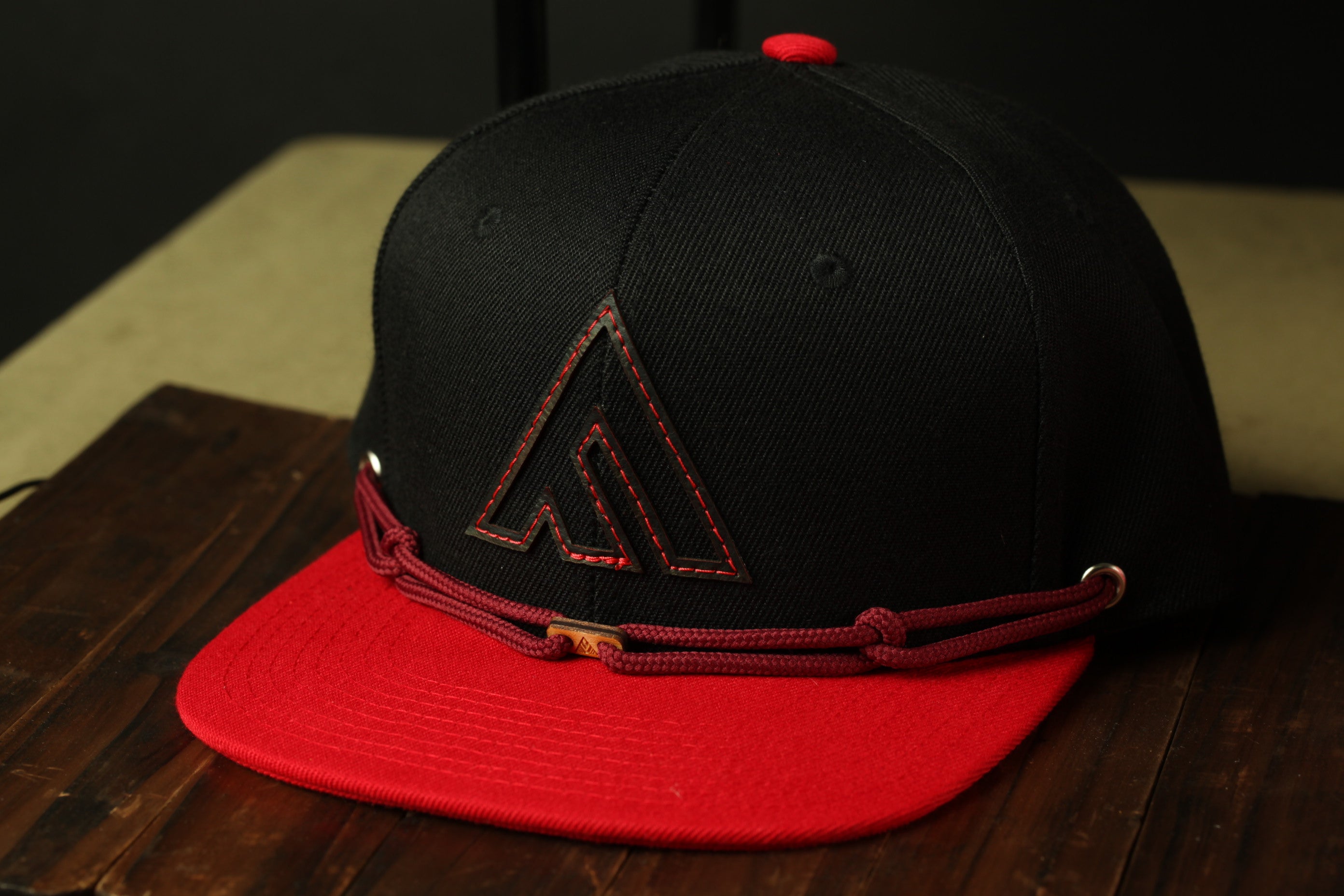A black baseball cap with a red brim and a red logo on the front, placed on a wooden surface.