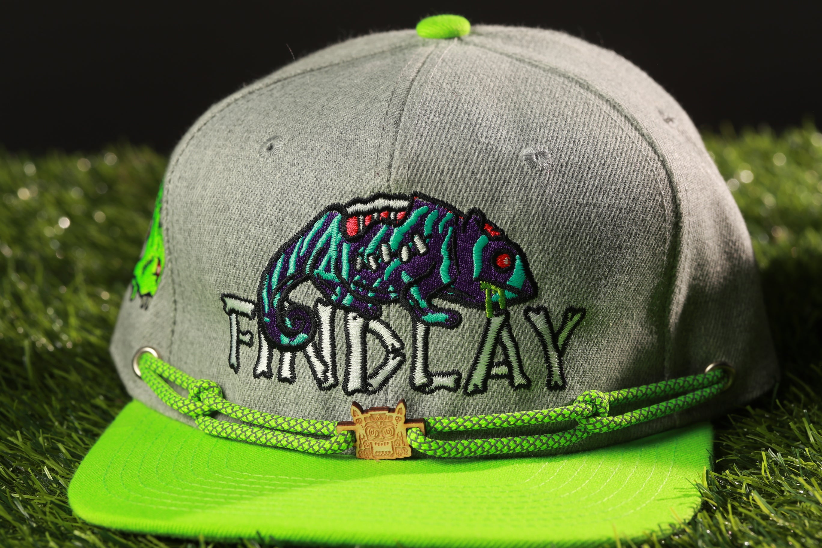 A grey baseball cap with a green brim and a chameleon design on the front, sitting on a grassy surface.