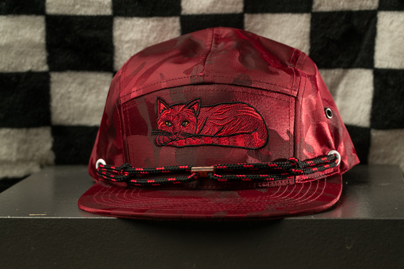 A red camouflage baseball cap with a cat embroidered on it, sitting on a black surface against a black and white checkered background.