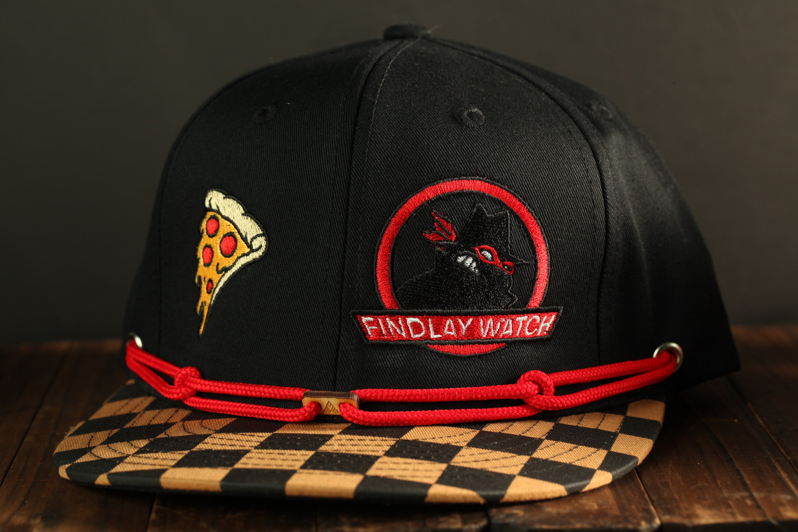 A black baseball cap with a red string, featuring a pizza slice and a logo for "Findlay Watch".