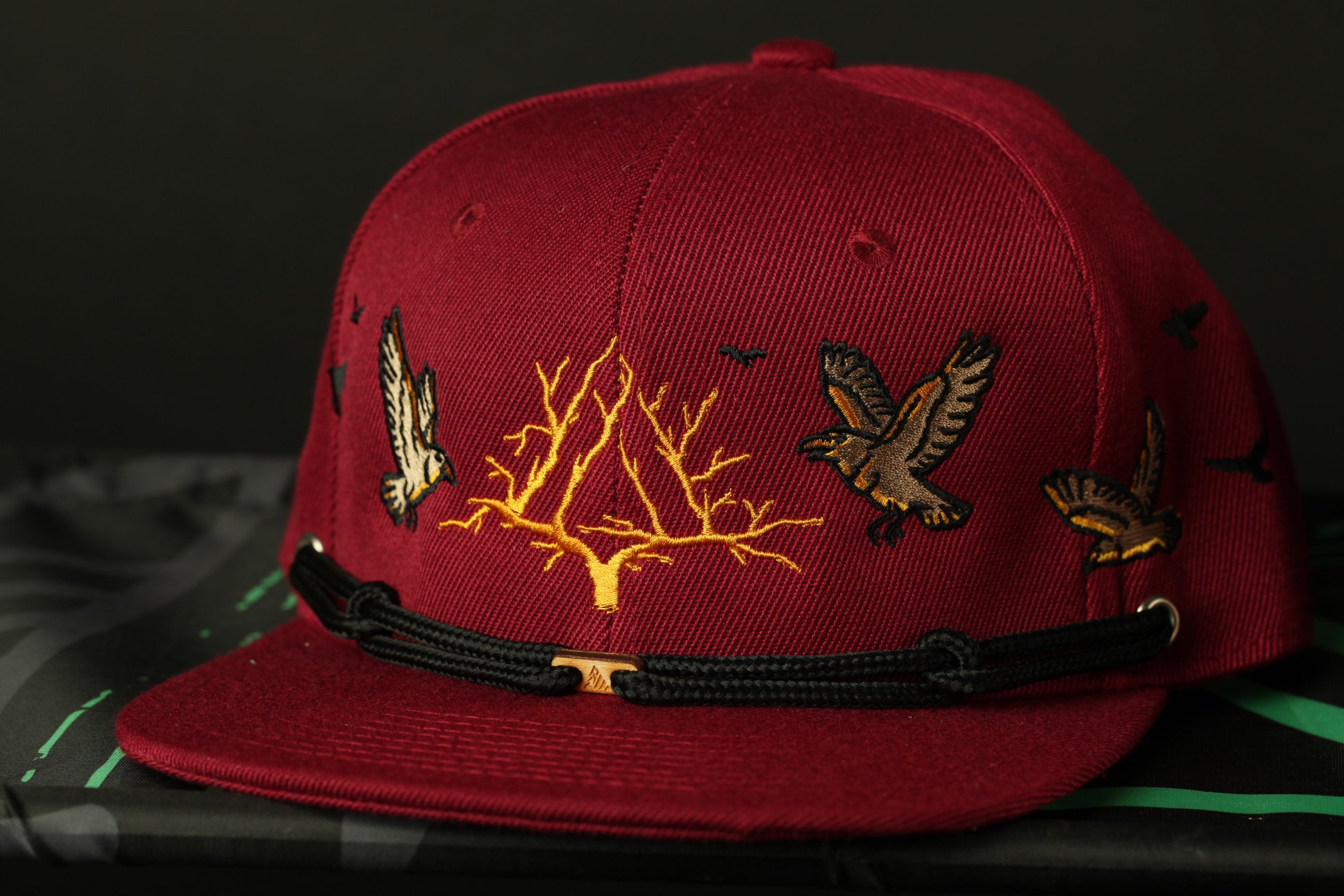 A red baseball cap with a black strap and gold embroidered designs of birds and a tree on the front.