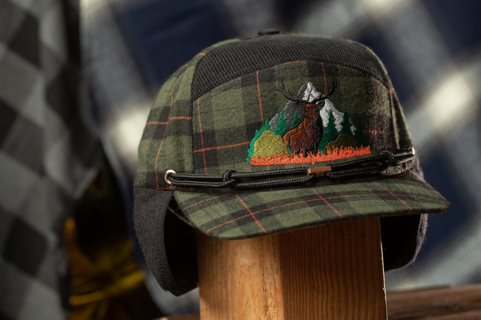 A plaid hat with a mountain and deer embroidered on it, displayed on a wooden stand.