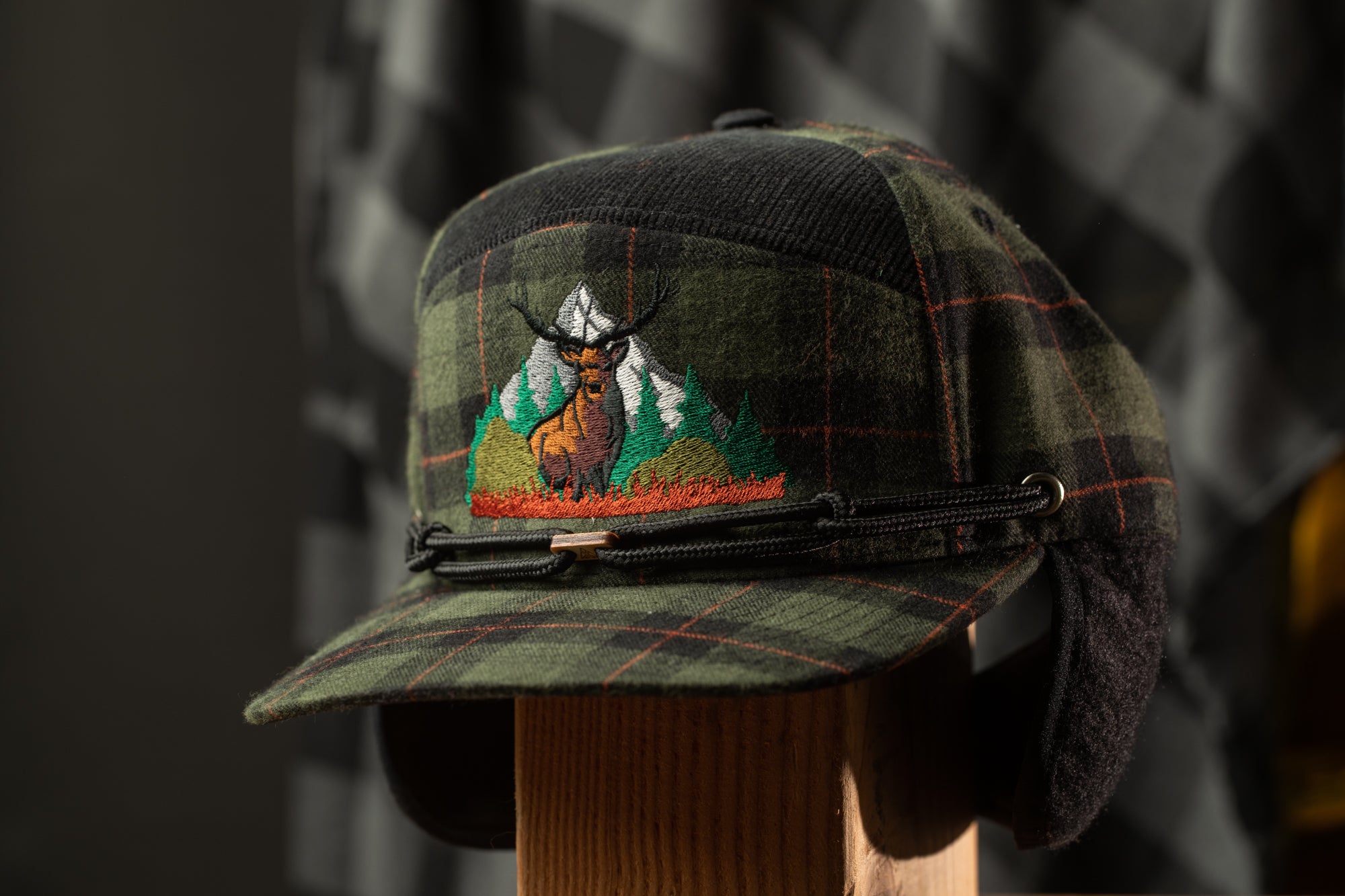A plaid baseball cap with an embroidered design of a deer standing on a mountain.