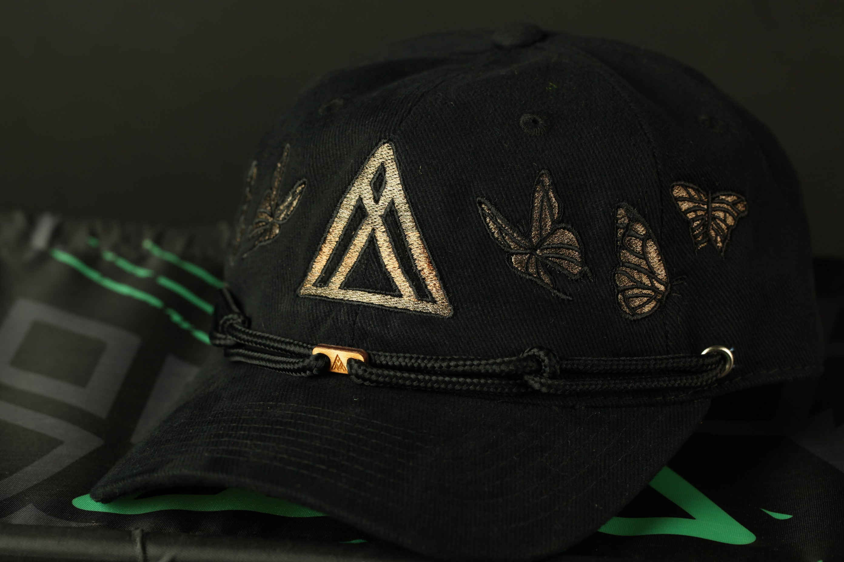 A black baseball cap with a gold triangle design and butterflies on the brim, resting on a black surface.