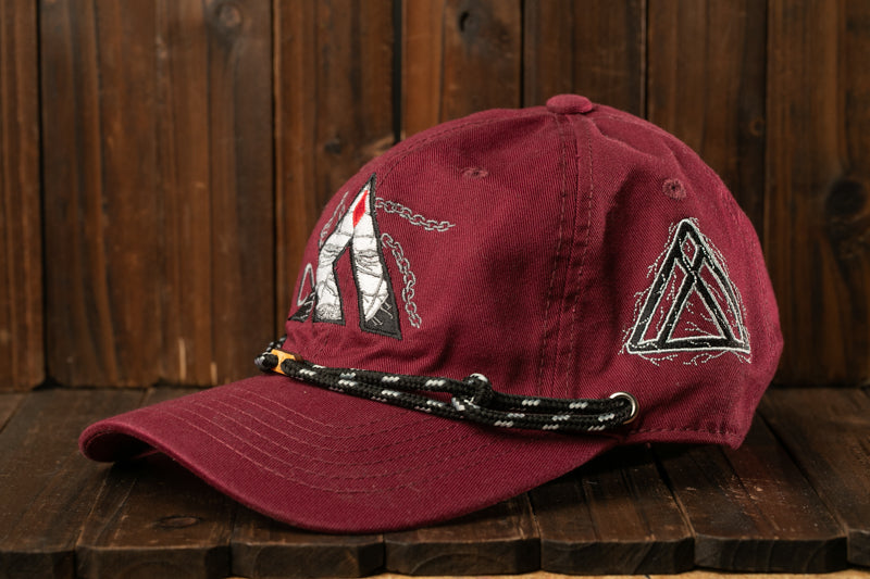 A maroon baseball cap with a black and white geometric design on the front and a black rope strap, placed on a wooden surface.