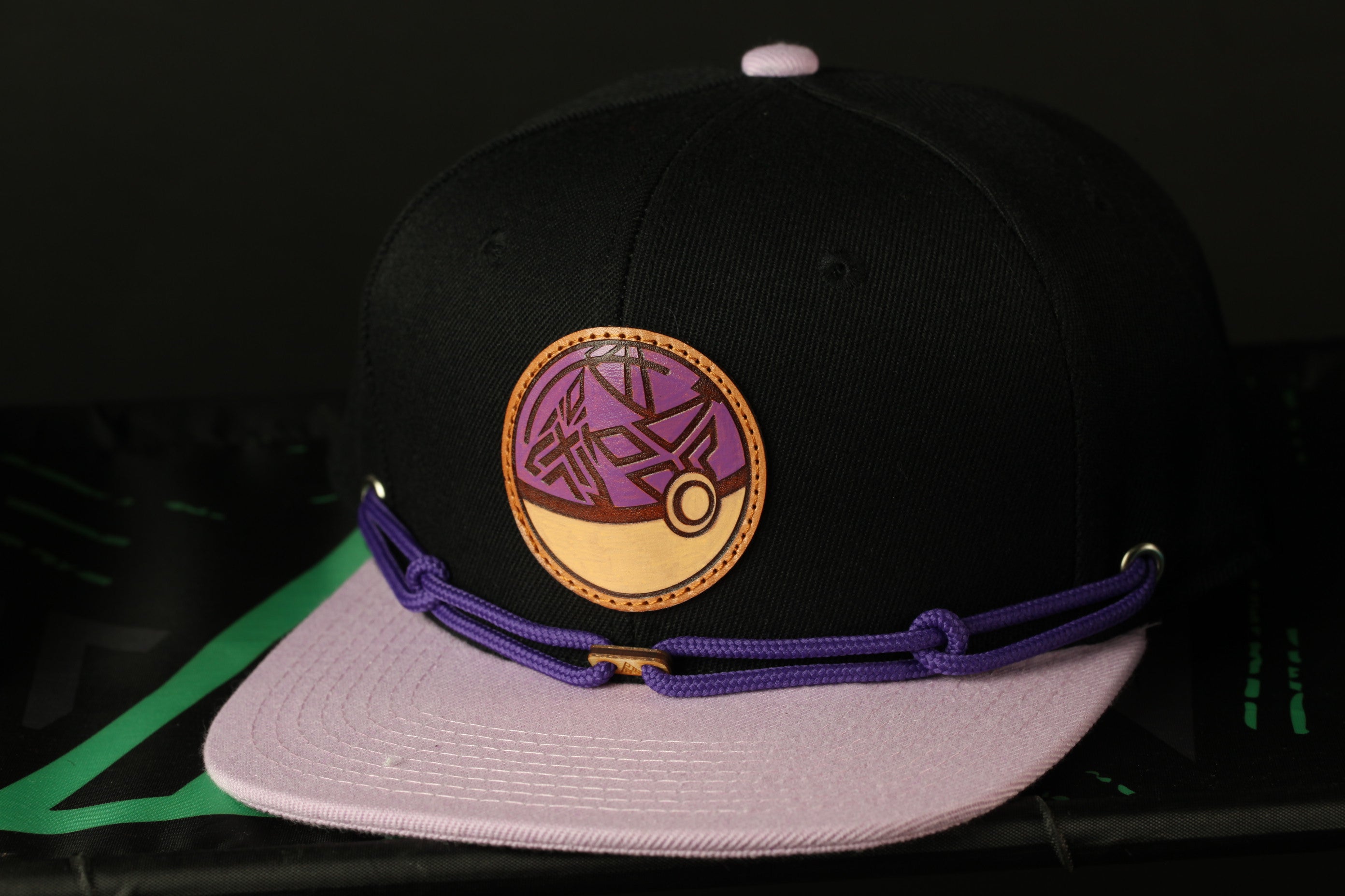 A black baseball cap with a purple and gold circular logo on the front, resting on a black surface.