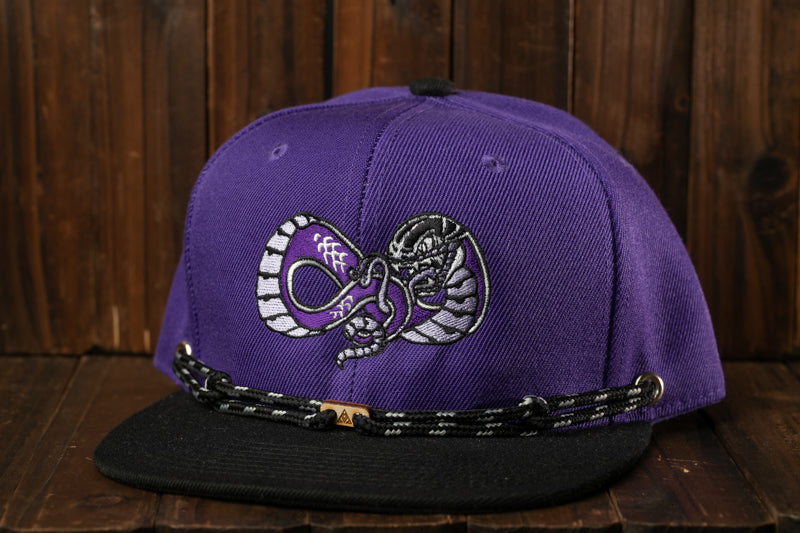 A purple baseball cap with a black brim and a black and white snake design on the front, sitting on a wooden surface.