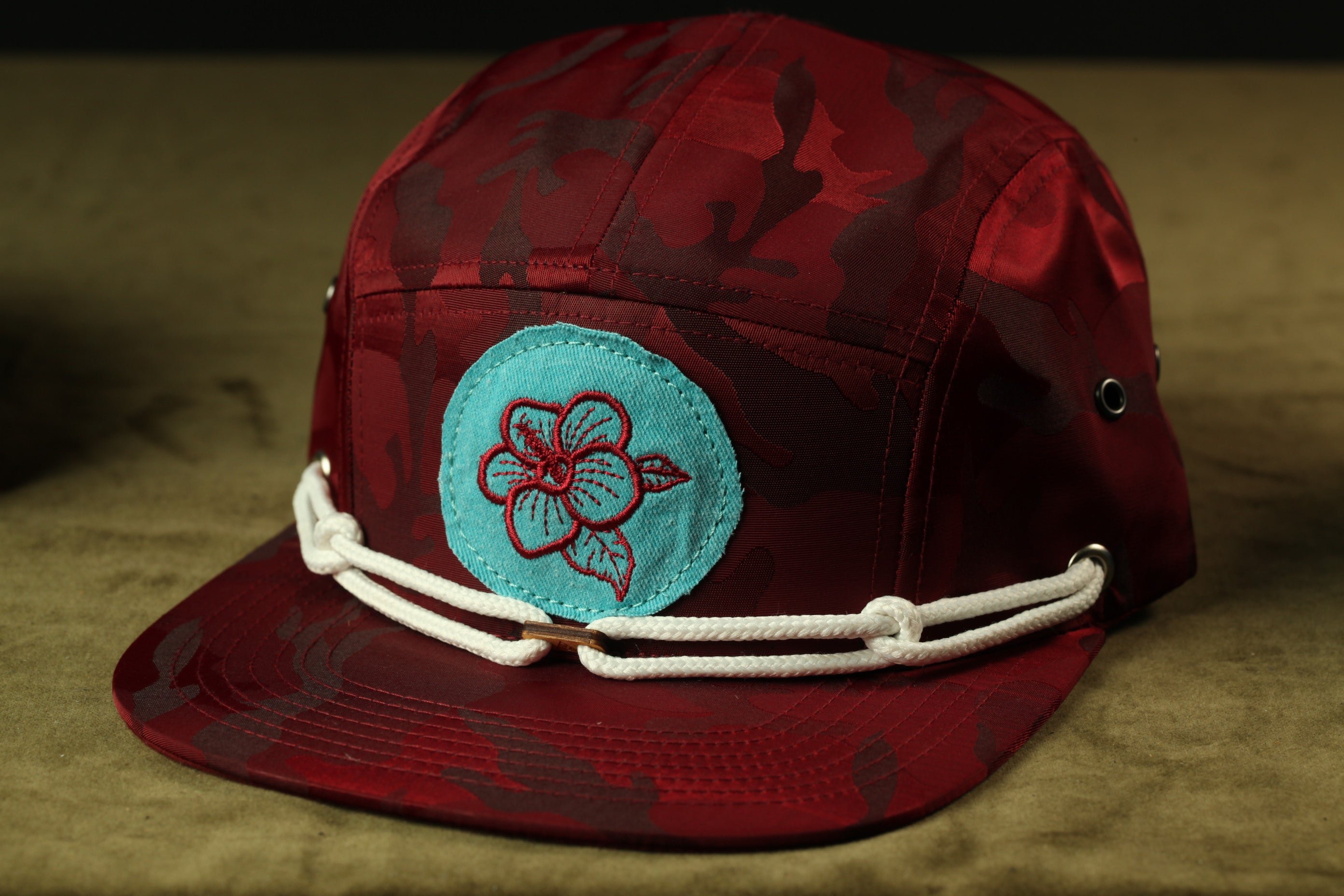 A maroon baseball cap with a blue flower patch and white rope strap.